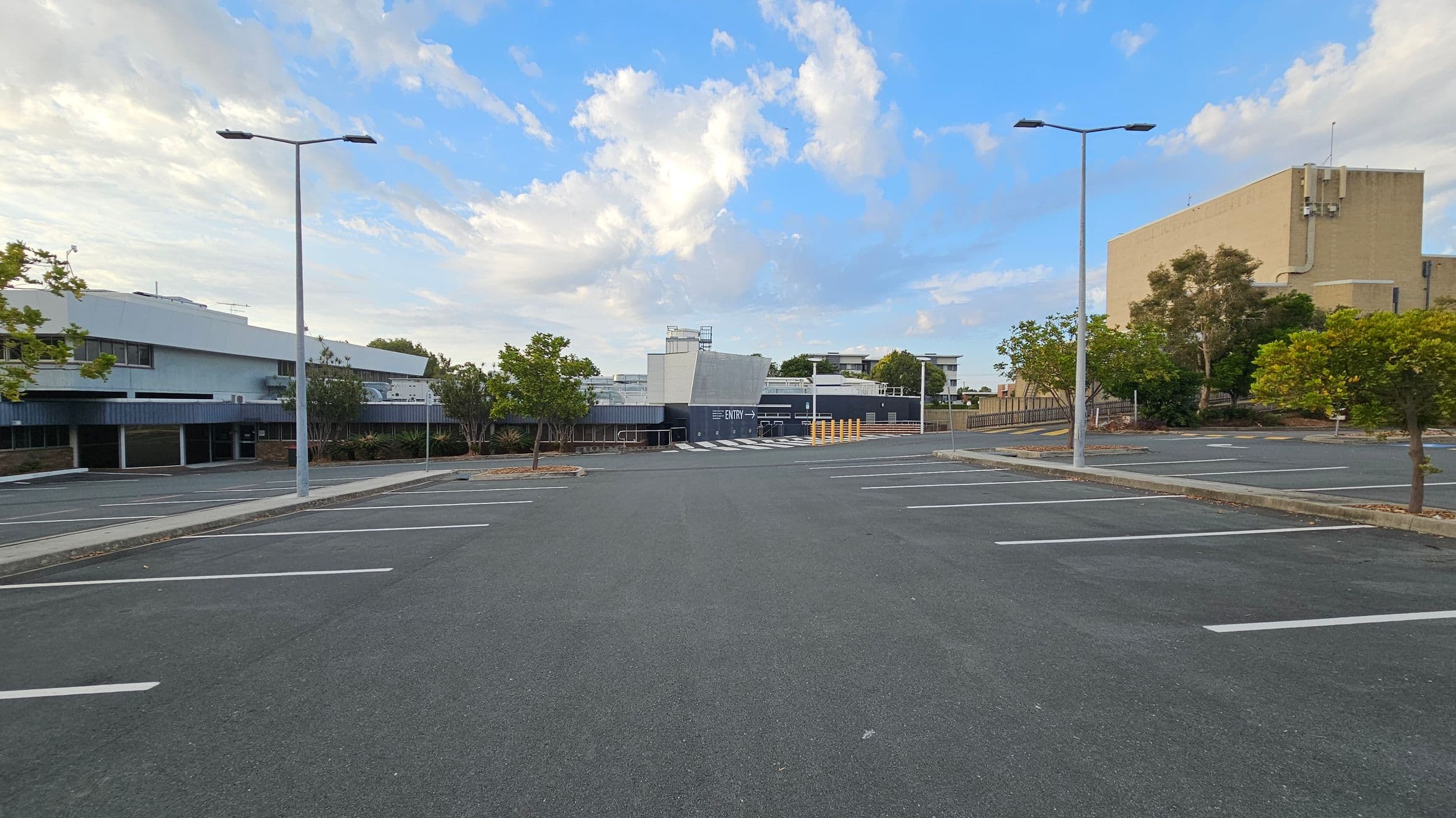 An empty car park with marked spaces and two tall street lamps. On the left, a modern building with a flat roof and large windows. In the centre, a structure with a sign indicating "ENTRY" and a pedestrian crossing. On the right, a large beige building with trees and shrubs around it. The ground is asphalt, and there are small trees planted along the edges of the car park.