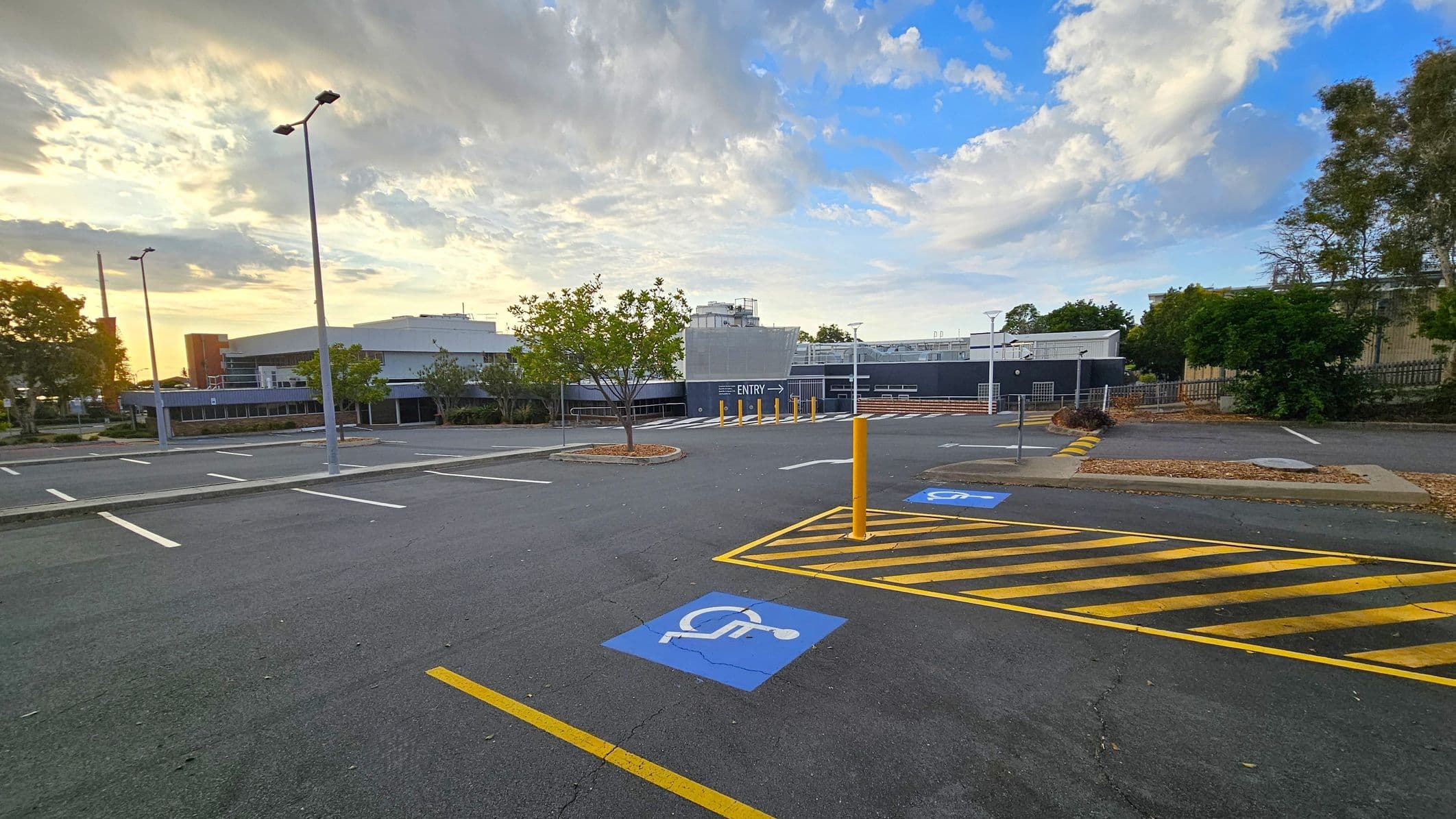 An empty car park with marked parking spaces, including a blue disabled parking spot in the foreground. Yellow and black striped lines are painted on the ground nearby. Several tall streetlights are positioned throughout the area. In the background, a building with a sign indicating "ENTRY" is visible, surrounded by trees and shrubs. The sky is partly cloudy with a mix of light and darker clouds.