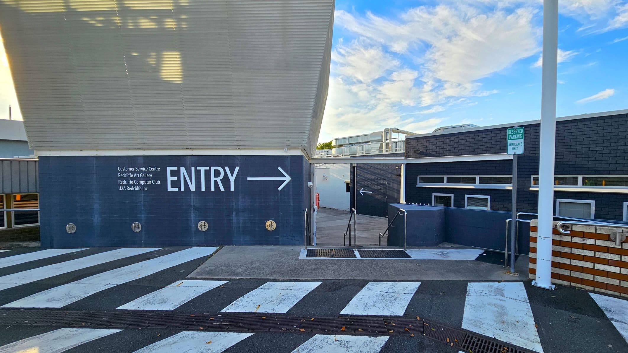 A building entrance with a large sign reading "ENTRY" and an arrow pointing right. The wall lists several facilities, including a customer service centre and art gallery. A zebra crossing is in the foreground, leading to a ramp with metal railings. A sign indicates reserved parking for ambulances. The building has a modern design with dark brick and metal elements. A white pole is visible on the right.