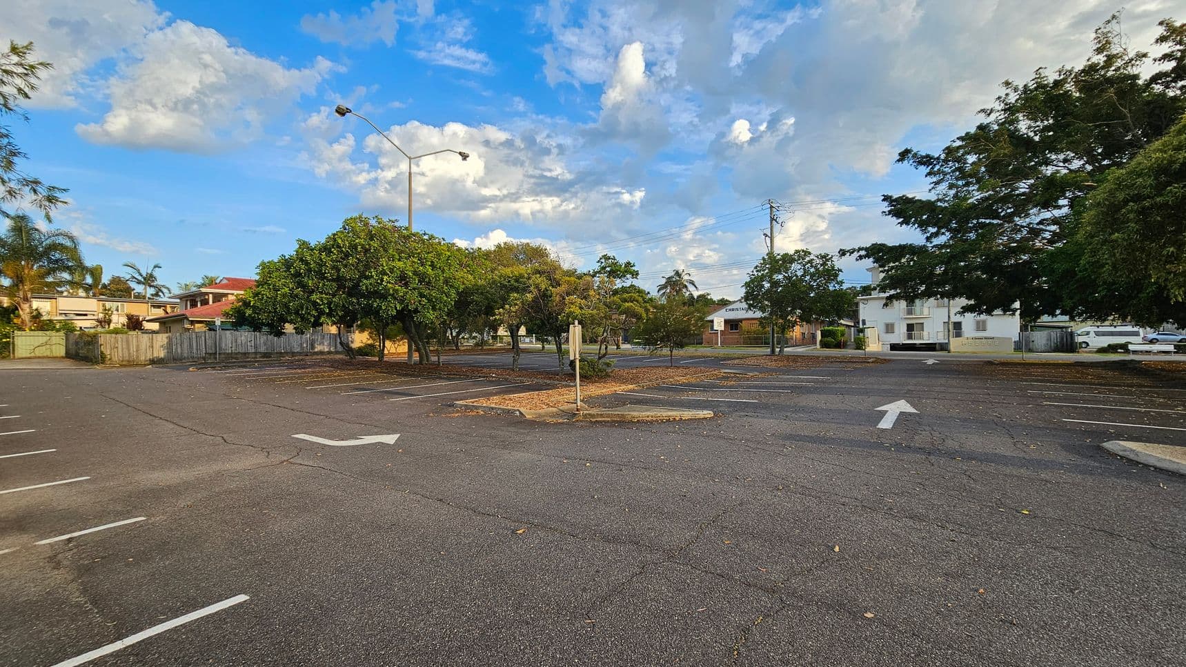 An empty car park with marked spaces and directional arrows on the asphalt. In the middle, there are several trees with green foliage, and a streetlamp is visible. To the left, a wooden fence and residential buildings with red roofs are seen. On the right, a white building and parked vehicles are present. The ground is scattered with dry leaves, and the sky is partly cloudy.