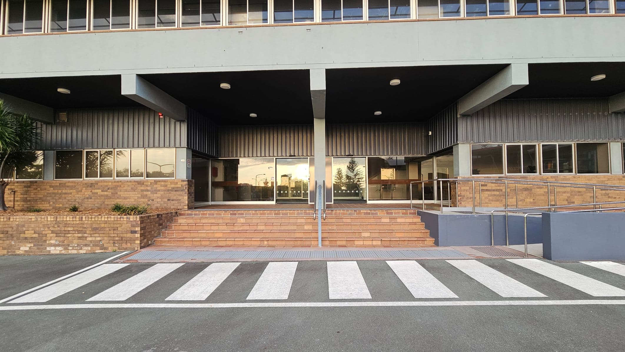 A building entrance with a series of steps leading up to glass doors. The facade features large windows above and brickwork below. A metal handrail is centred on the steps. To the right, a ramp with a metal railing provides accessibility. A zebra crossing is painted on the asphalt in the foreground, leading directly to the entrance. The ceiling above the entrance is dark with several round lights.