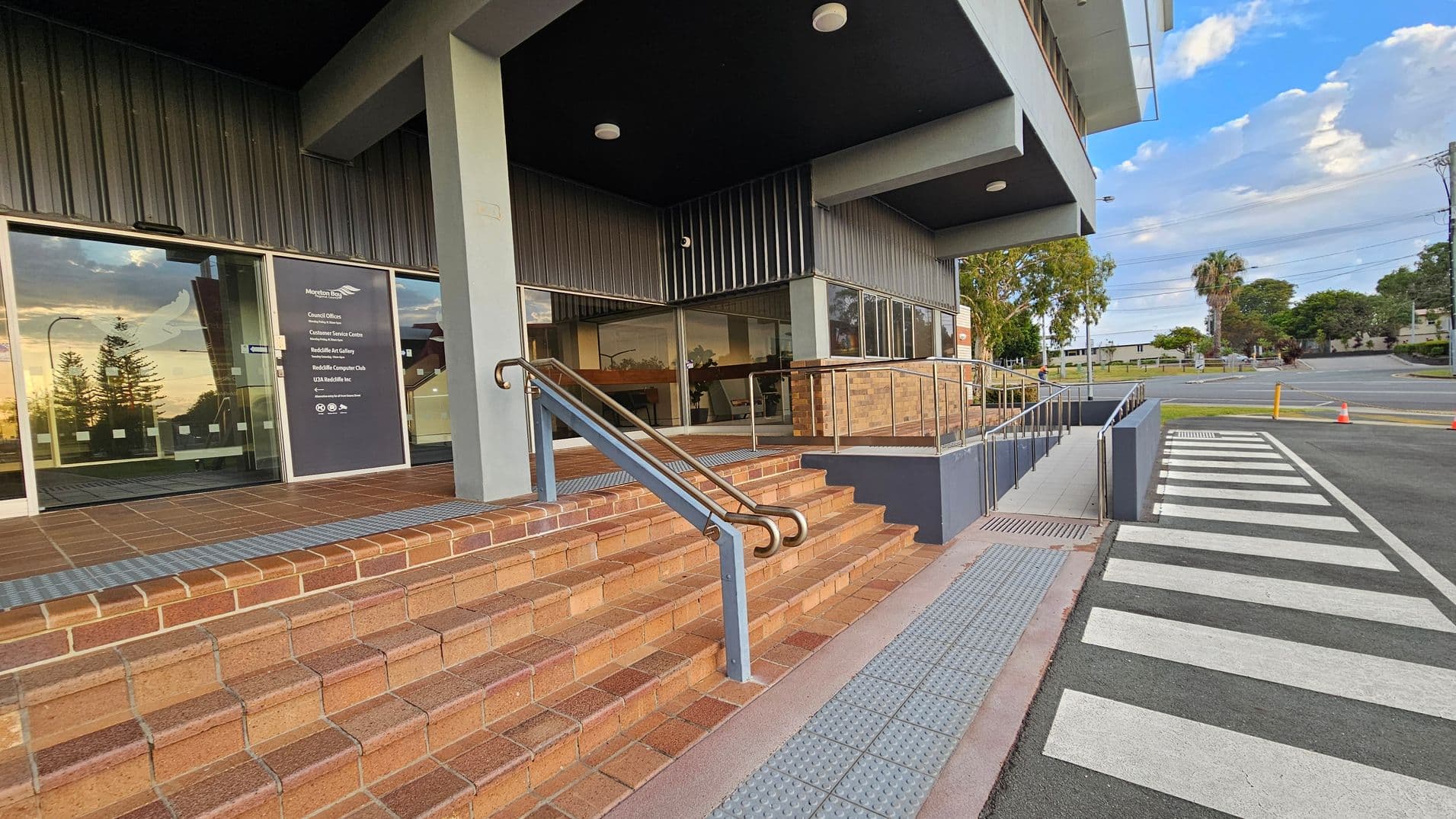 A building entrance with brown brick steps and a metal handrail on the left. Glass doors with signage are visible, reflecting the surroundings. To the right, a ramp with a railing leads to the entrance, adjacent to a zebra crossing on the road. The ceiling is dark with round lights. In the background, there are trees and a clear sky.