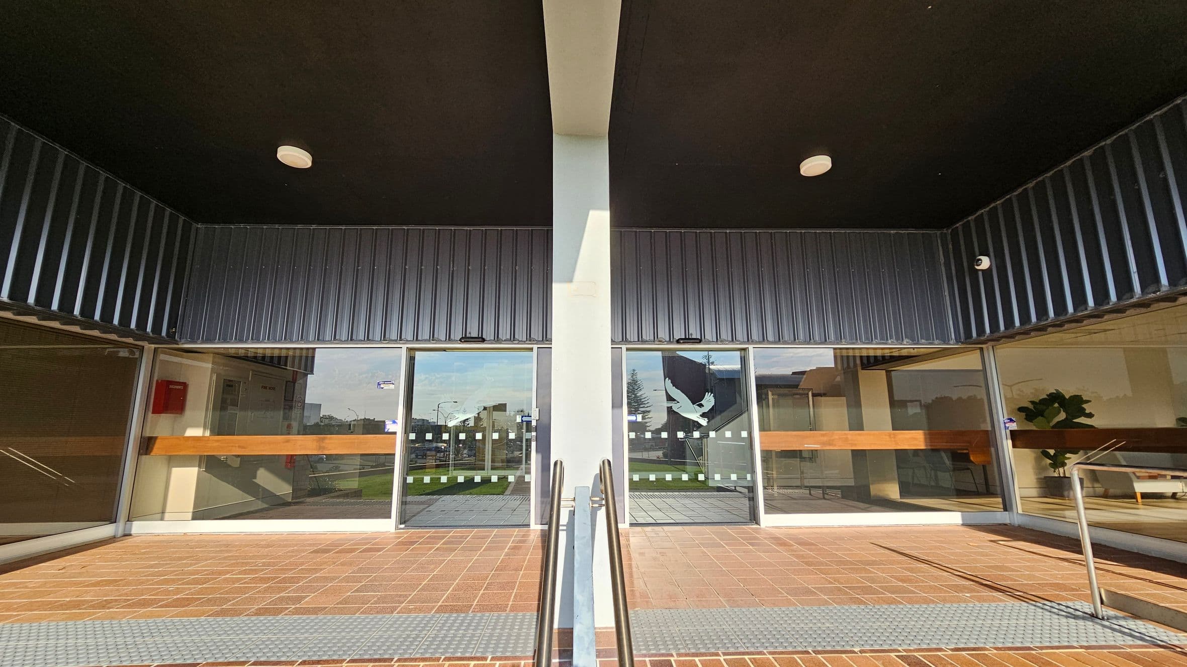 A symmetrical entrance with two glass doors, each featuring a bird design. The doors are flanked by large glass panels. A central pillar divides the entrance, with metal railings leading up to it. The floor is tiled in a reddish-brown pattern, and the ceiling is dark with circular lights. Inside, there are visible plants and furniture.