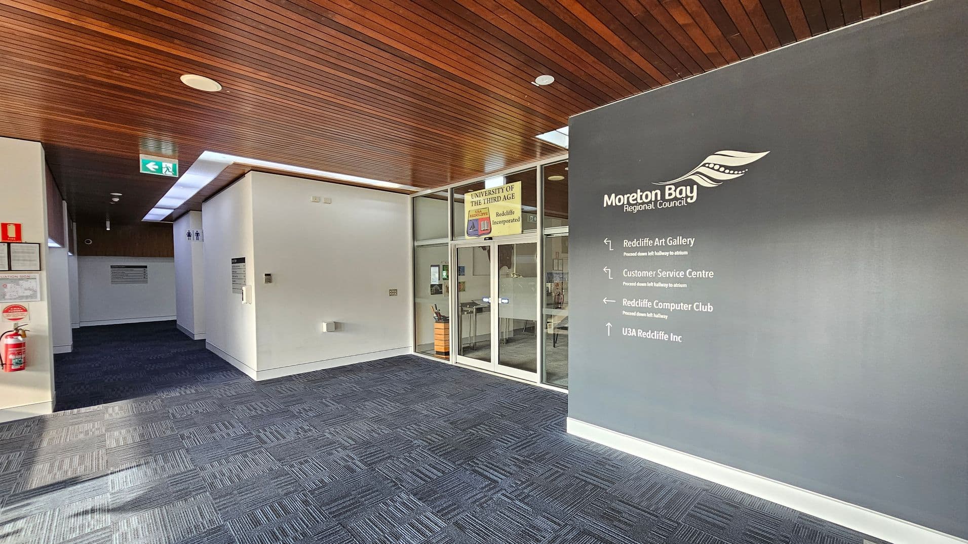 A hallway with a dark patterned carpet and a wooden panelled ceiling. On the right, a grey wall displays the Moreton Bay Regional Council logo and directional signs for various facilities. On the left, a fire extinguisher is mounted on the wall. In the centre, glass doors lead to the University of the Third Age, with a sign above. An exit sign is visible on the ceiling, and there are restroom signs on the left wall.