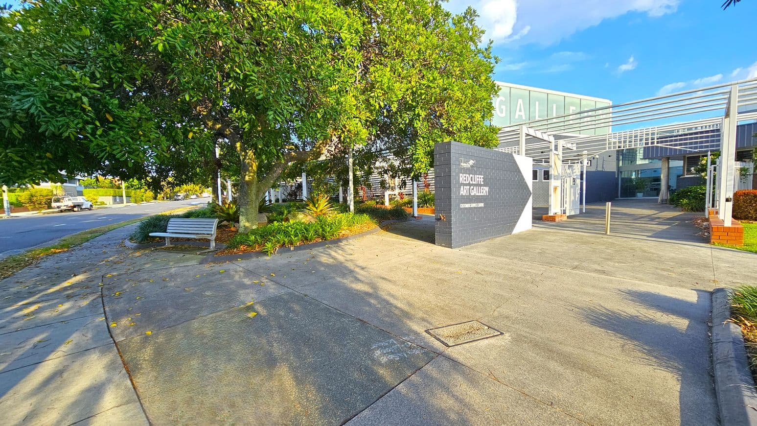 A paved pathway leads to the entrance of a building with a sign reading "Redcliffe Art Gallery" on a dark brick wall. On the left, a large tree with dense green foliage provides shade over a white bench. The road runs parallel to the path, with a parked vehicle visible in the distance. The building features a modern design with metal and glass elements, and there are landscaped plants around the entrance. Shadows from the tree and structure are cast on the ground.