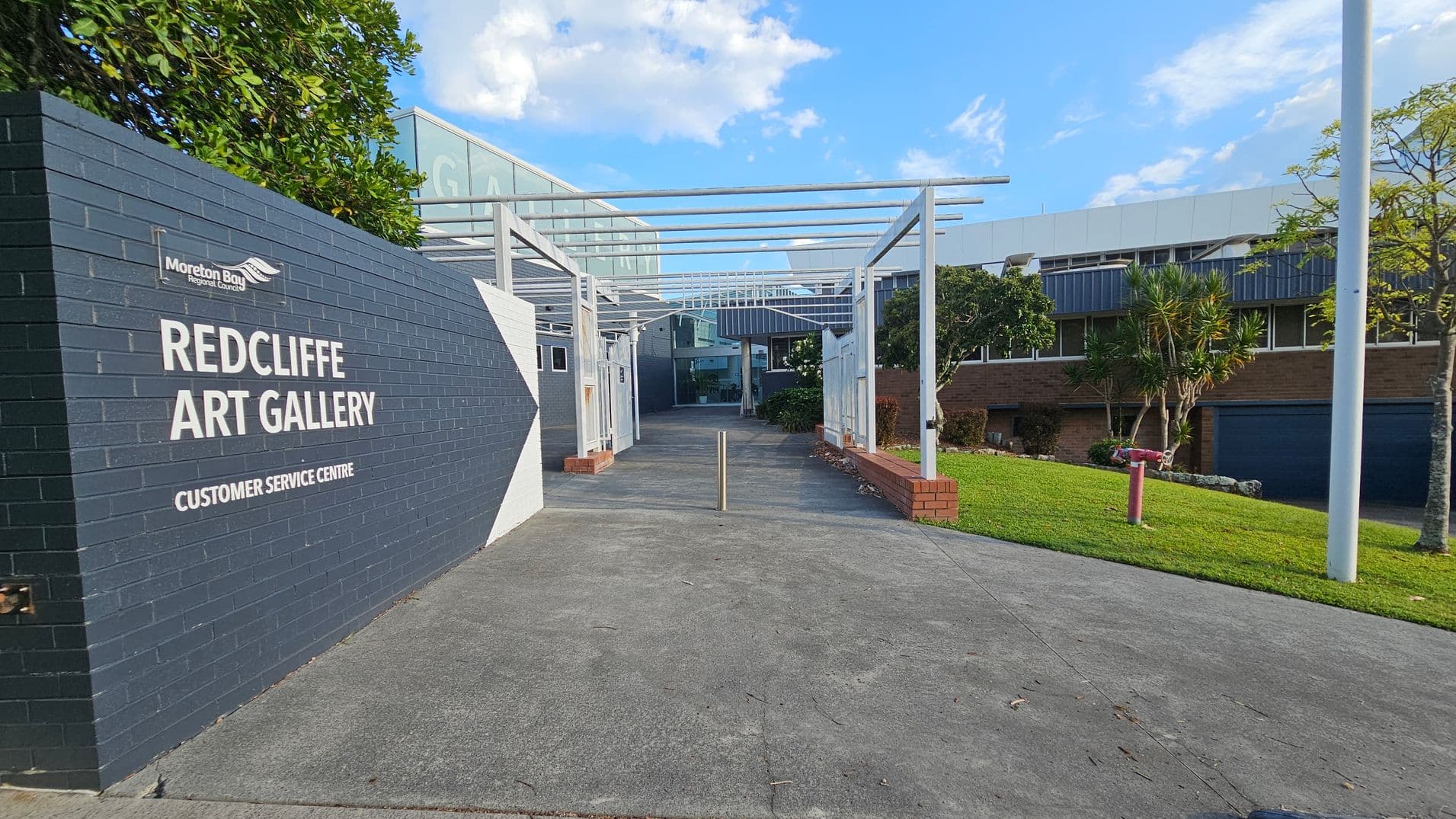 A pathway leads to the entrance of the Redcliffe Art Gallery, with a dark brick wall on the left displaying the gallery's name and a logo. The path is flanked by white metal structures and bordered by low brick walls. On the right, there is a grassy area with small trees and a red fire hydrant. The building in the background has a modern design with large windows and a mix of brick and metal elements. The sky is partly cloudy.