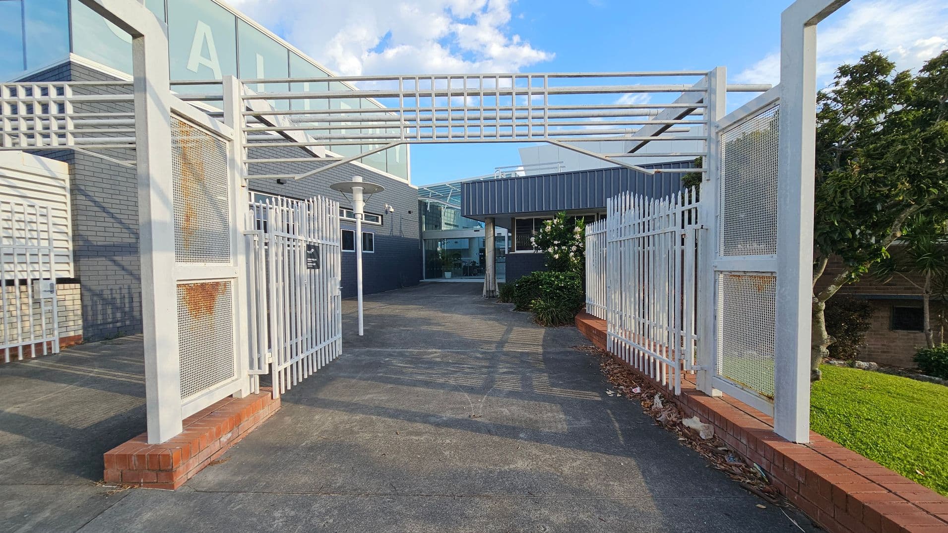 A pathway leads through a white metal gate with mesh panels, some showing rust. The path is bordered by a low red brick wall. On the left, a grey brick building with a large glass section is visible. On the right, a garden with green shrubs and a tree. The ground is concrete, and there are shadows from the gate. A modern lamp post stands near the entrance.