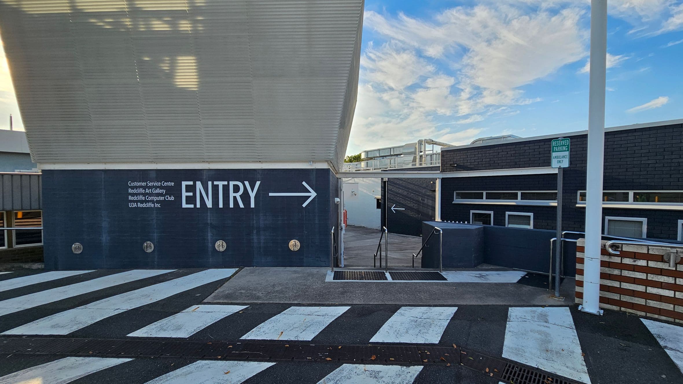 A building entrance with a large "ENTRY" sign and an arrow pointing right. The wall lists several facilities, including a customer service centre and art gallery. The foreground features a striped pedestrian crossing. On the right, a sign indicates reserved parking for ambulances. The building has a dark exterior with small windows, and there are metal railings leading to the entrance. The ceiling is slanted and white.