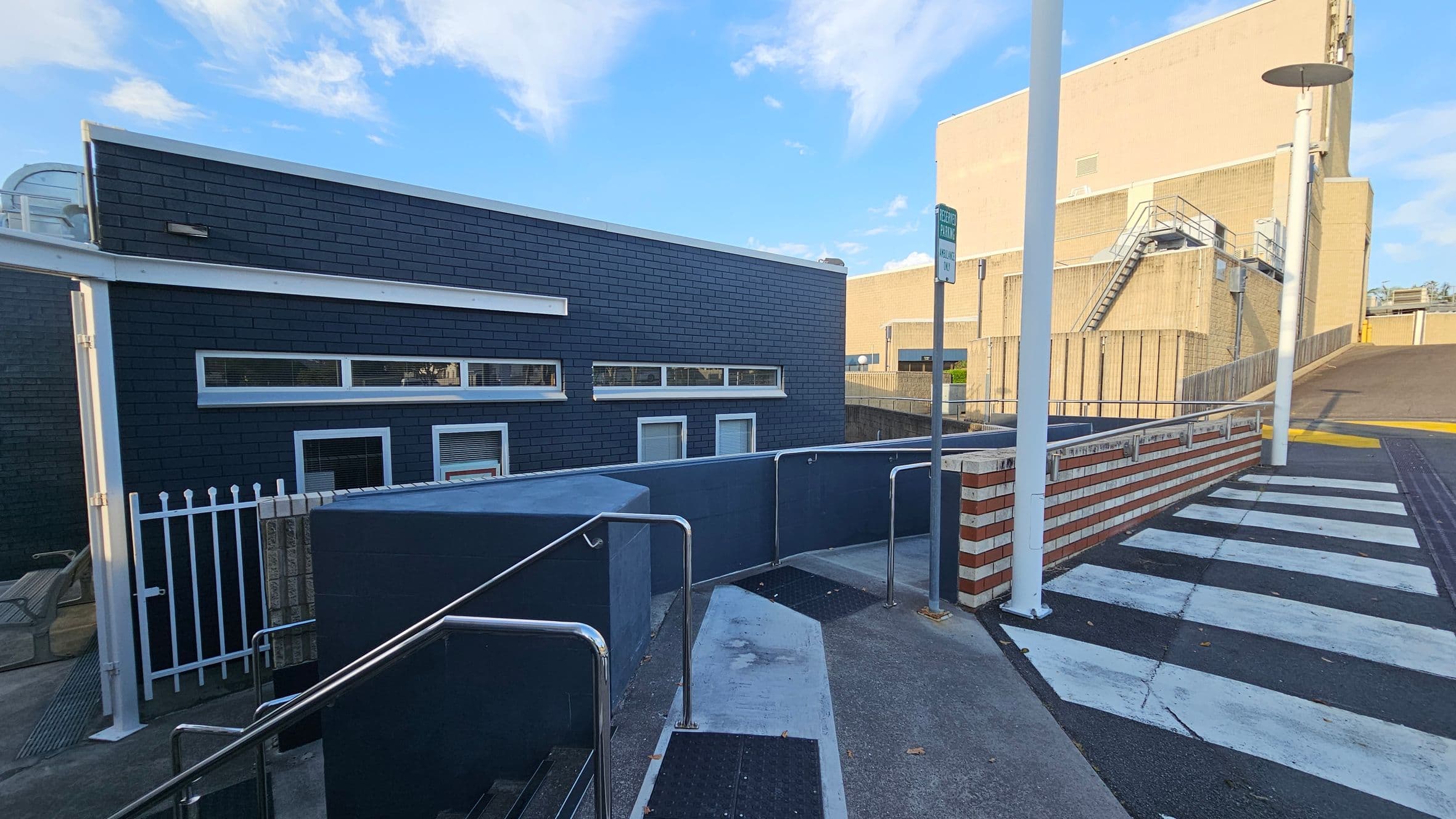A modern building with dark blue brick walls and narrow horizontal windows is on the left. In front, a white metal fence and a ramp with metal railings lead up to a pedestrian crossing marked with white stripes. On the right, a tall beige building with external stairs is visible. A streetlamp and a signpost are near the crossing. The ground is paved with concrete and asphalt, and the sky is clear with a few clouds.
