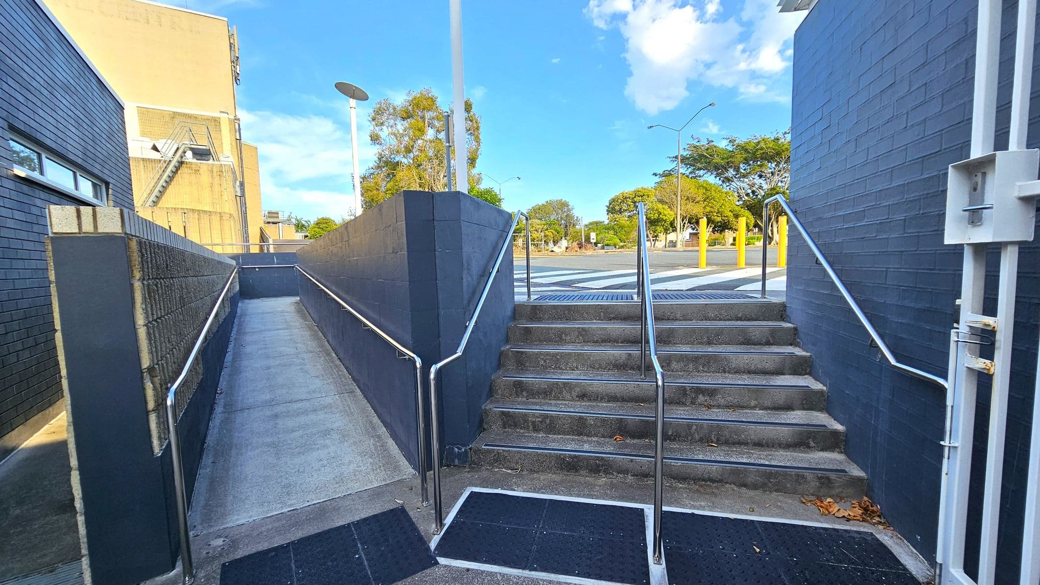 A concrete ramp with metal railings is on the left, leading upwards between dark brick walls. On the right, a set of concrete steps with similar railings ascends to the same level. In the background, a street with a zebra crossing and yellow bollards is visible, along with trees and a building with a beige facade. The sky is clear with a few clouds.