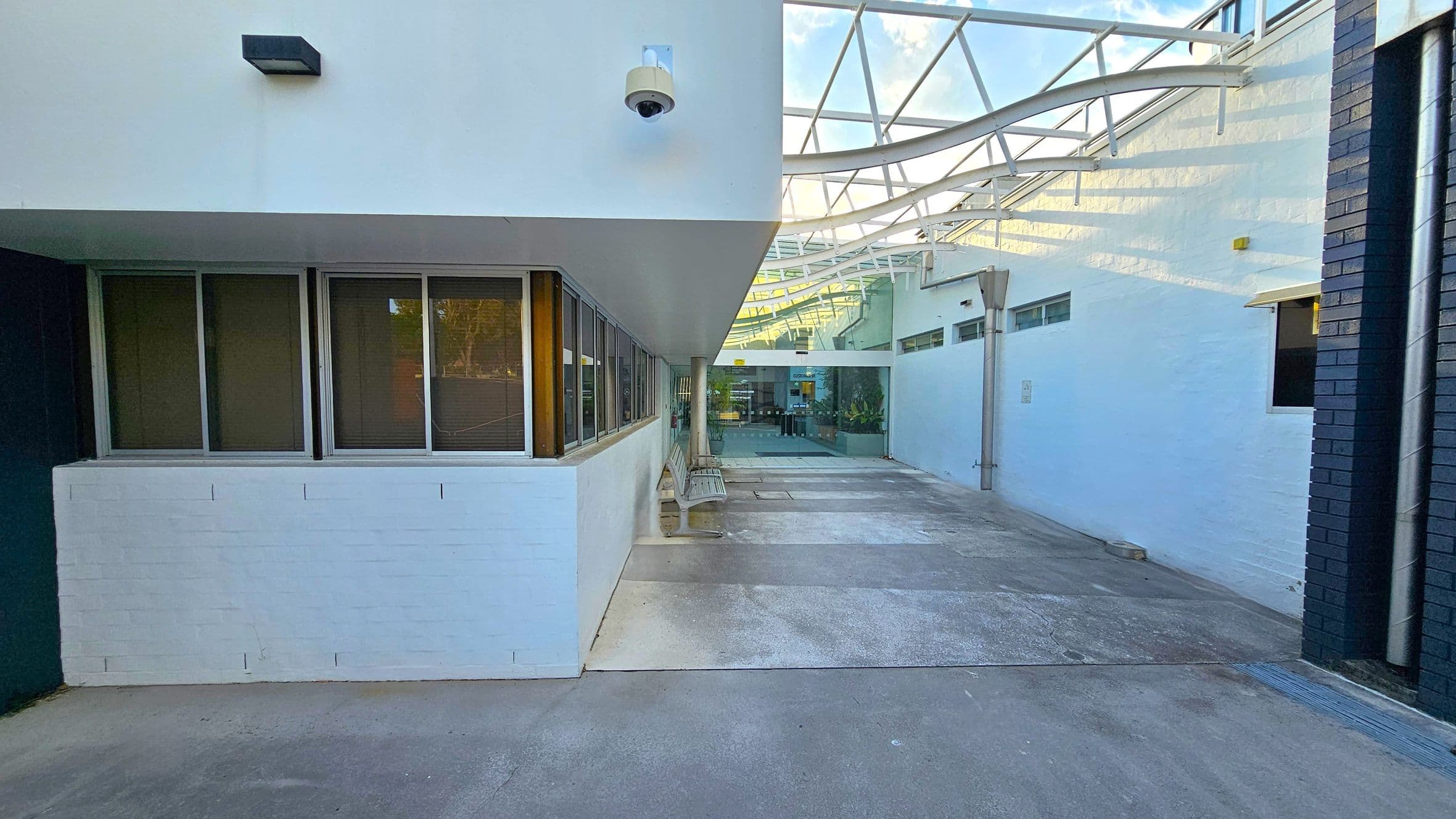 A walkway with a concrete floor and a white ceiling featuring a security camera. On the left, there are windows with metal frames and a bench beneath them. The right side has a white wall with a black brick corner. The background shows a glass entrance with plants and a metal framework above, allowing natural light to filter through.