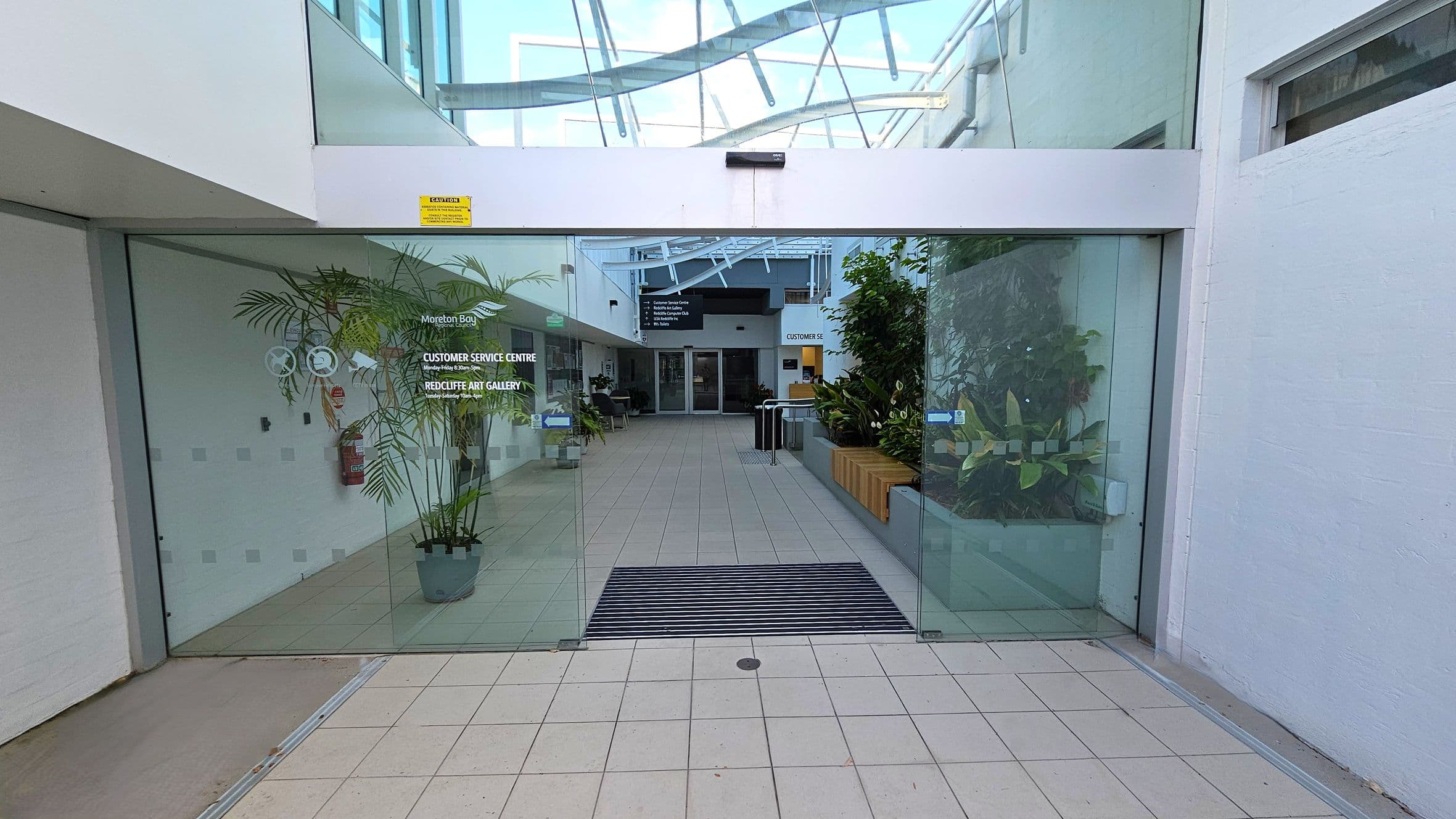 Glass doors with signage for a customer service centre and art gallery. Inside, a tiled floor leads to a hallway with potted plants on both sides. On the left, a fire extinguisher is mounted on the wall. On the right, a wooden bench is surrounded by more plants. The ceiling is made of glass panels with metal beams. A sign in the background provides directions.