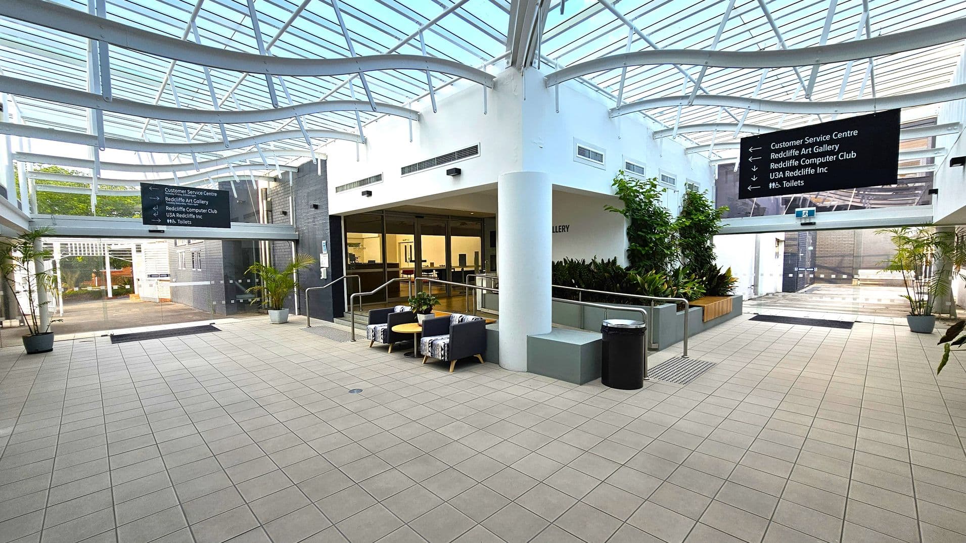 A spacious indoor area with a tiled floor and a glass ceiling featuring a metal framework. In the centre, there are two cushioned chairs and a small round table. To the right, a white column and a black bin are near a wall with greenery. On the left, a sign directs to various locations, and there are potted plants. The background shows glass doors and a pathway leading outside.