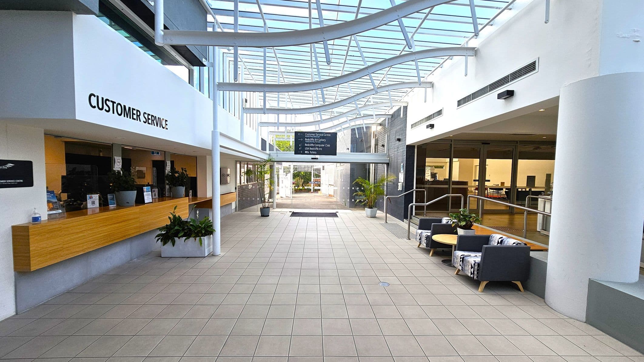 A spacious indoor area with a tiled floor and a glass ceiling featuring curved beams. On the left, there's a customer service counter with a wooden surface and potted plants. On the right, a seating area with patterned chairs and a round table is visible. In the centre, a signboard hangs above, and potted plants are placed near the entrance. The background shows an open doorway leading outside.