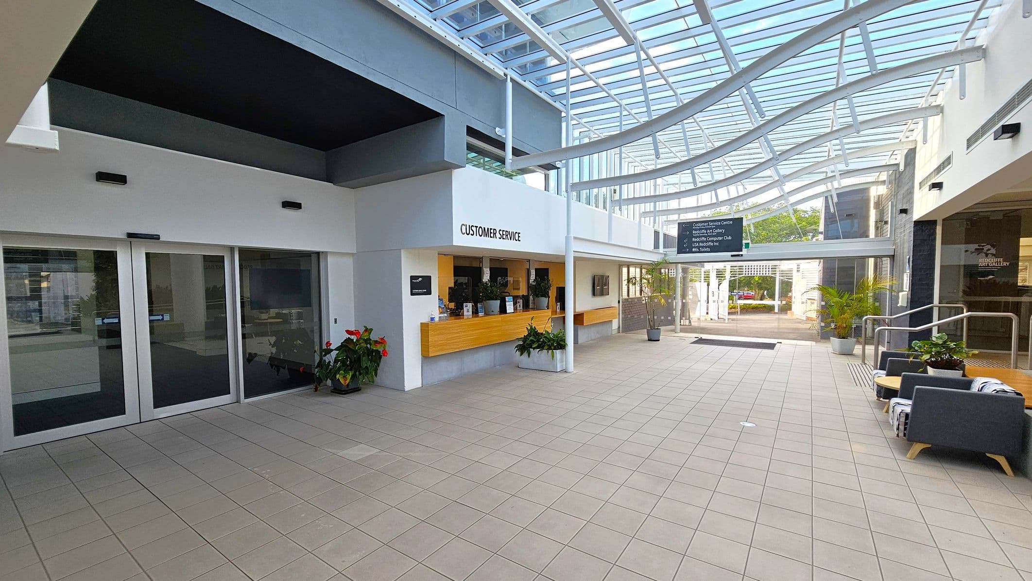 A spacious indoor area with a tiled floor and a glass ceiling. On the left, there are large glass doors and a customer service counter with a wooden front and potted plants. On the right, there are grey sofas with small tables and more potted plants. In the background, a sign hangs from the ceiling, and there is an entrance leading outside. The area is well-lit with natural light streaming through the glass ceiling.