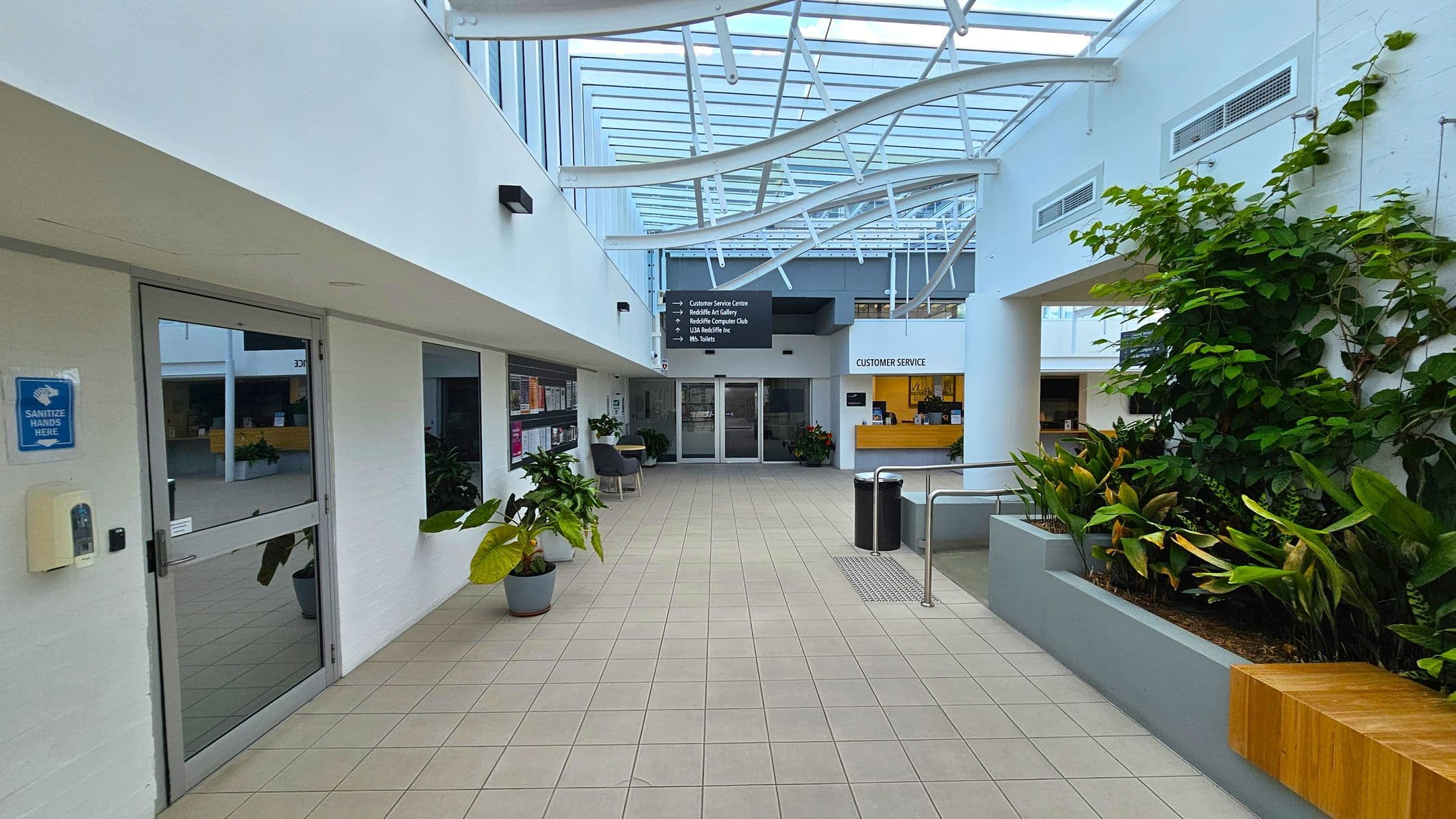 A spacious indoor corridor with a tiled floor and a glass ceiling featuring white structural beams. On the left, there's a glass door with a hand sanitiser dispenser and a sign. Potted plants and chairs line the wall. On the right, a raised planter with lush green plants and a wooden bench. In the centre, a customer service desk with a yellow backdrop. Overhead signs direct to various facilities.