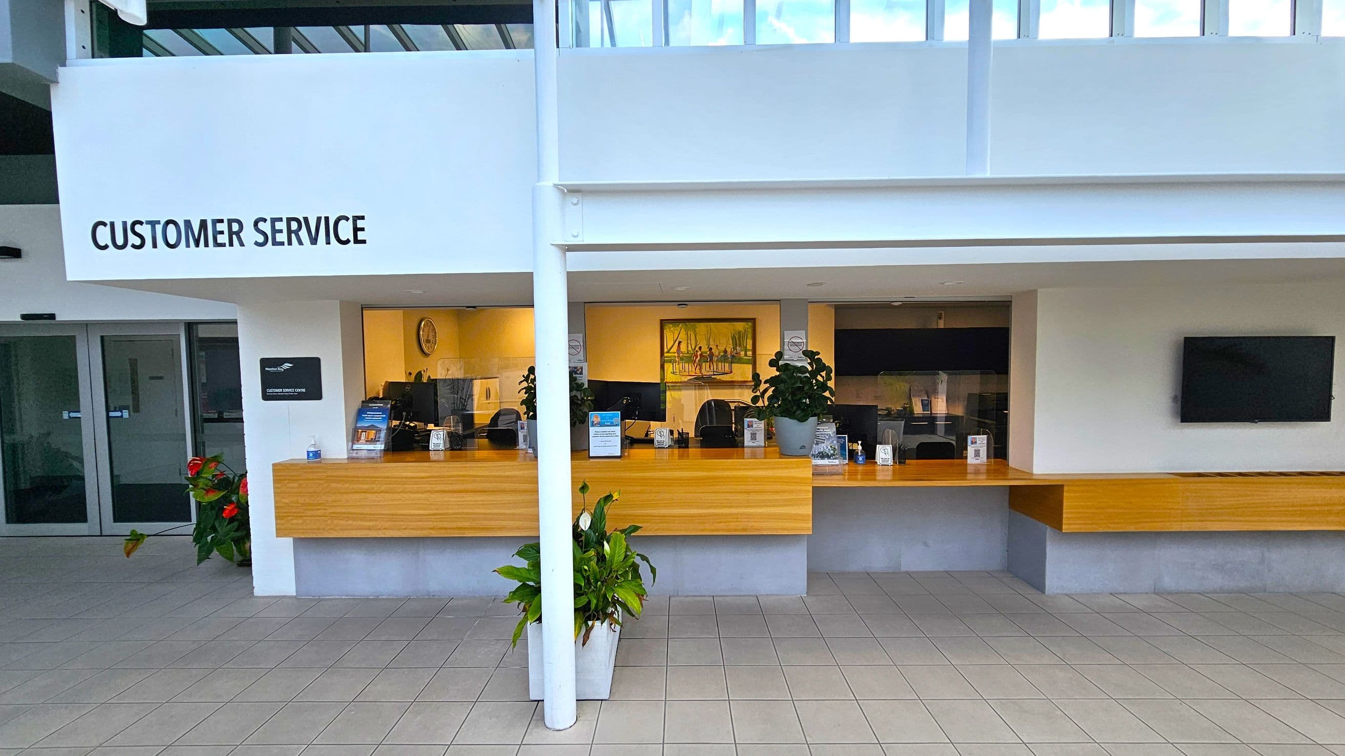 Customer service area with a wooden counter spanning the width. Behind the counter are several workstations with computers and potted plants. A painting and a clock are mounted on the wall. To the left, a sign reads "Customer Service" above a doorway with glass doors. On the right, a flat-screen TV is mounted on the wall. The floor is tiled, and there are potted plants in front of the counter. The ceiling is white with a skylight above.