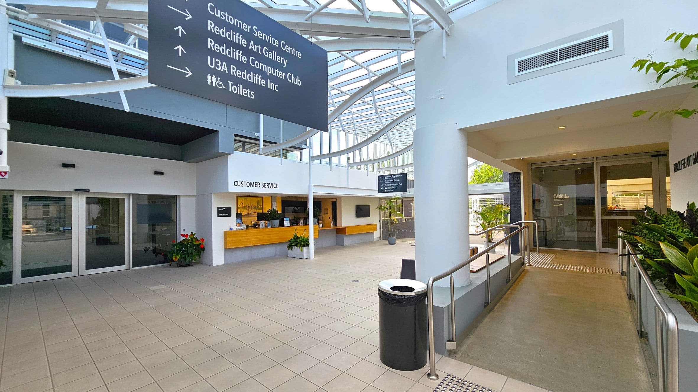 A spacious indoor area with a tiled floor and a high, glass-panelled ceiling. On the left, there are large glass doors and a customer service desk with a wooden counter. Above, a sign directs to various locations. On the right, a ramp leads to a glass door entrance, with plants lining the side. A black bin is positioned near the ramp. The area is well-lit with natural light streaming in through the ceiling.
