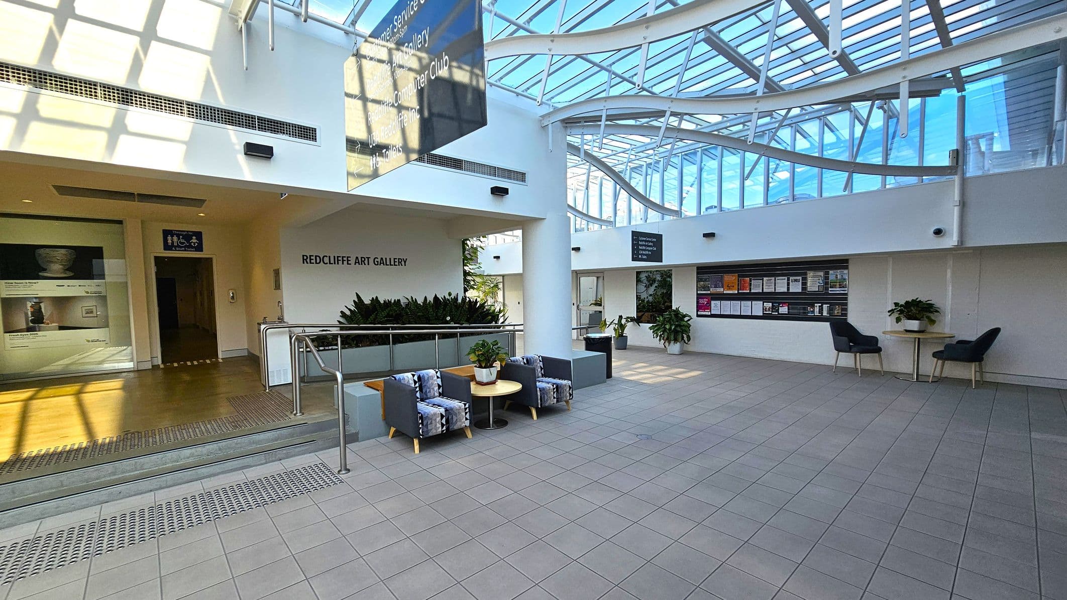 A spacious indoor area with a tiled floor and a glass ceiling allowing natural light. On the left, there's an entrance to the Redcliffe Art Gallery, with a sign above and a display of art posters. In the centre, a seating area with two patterned chairs and a small round table. On the right, a wall with notice boards and two chairs with a table. Potted plants are placed around the space, adding greenery. The ceiling features a modern, curved design with metal beams.
