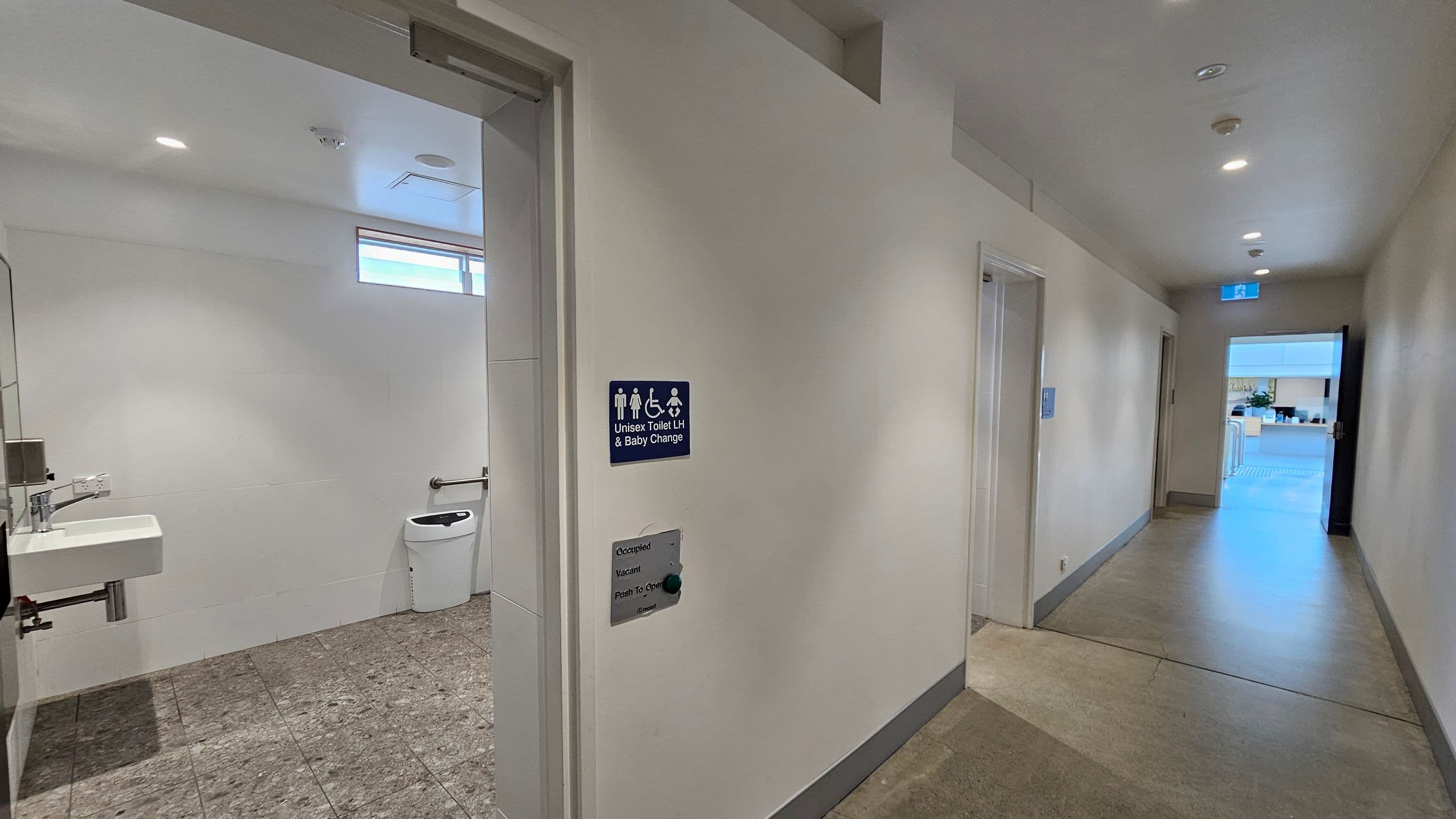 A hallway with a unisex toilet on the left, featuring a sign indicating accessibility and baby changing facilities. Inside, there's a sink and a toilet with a grab bar. The floor is tiled, and the walls are light-coloured. The ceiling has recessed lighting. On the right, a corridor leads to an open door with a view of an outdoor area. The floor transitions from tiles to a smooth surface in the hallway.