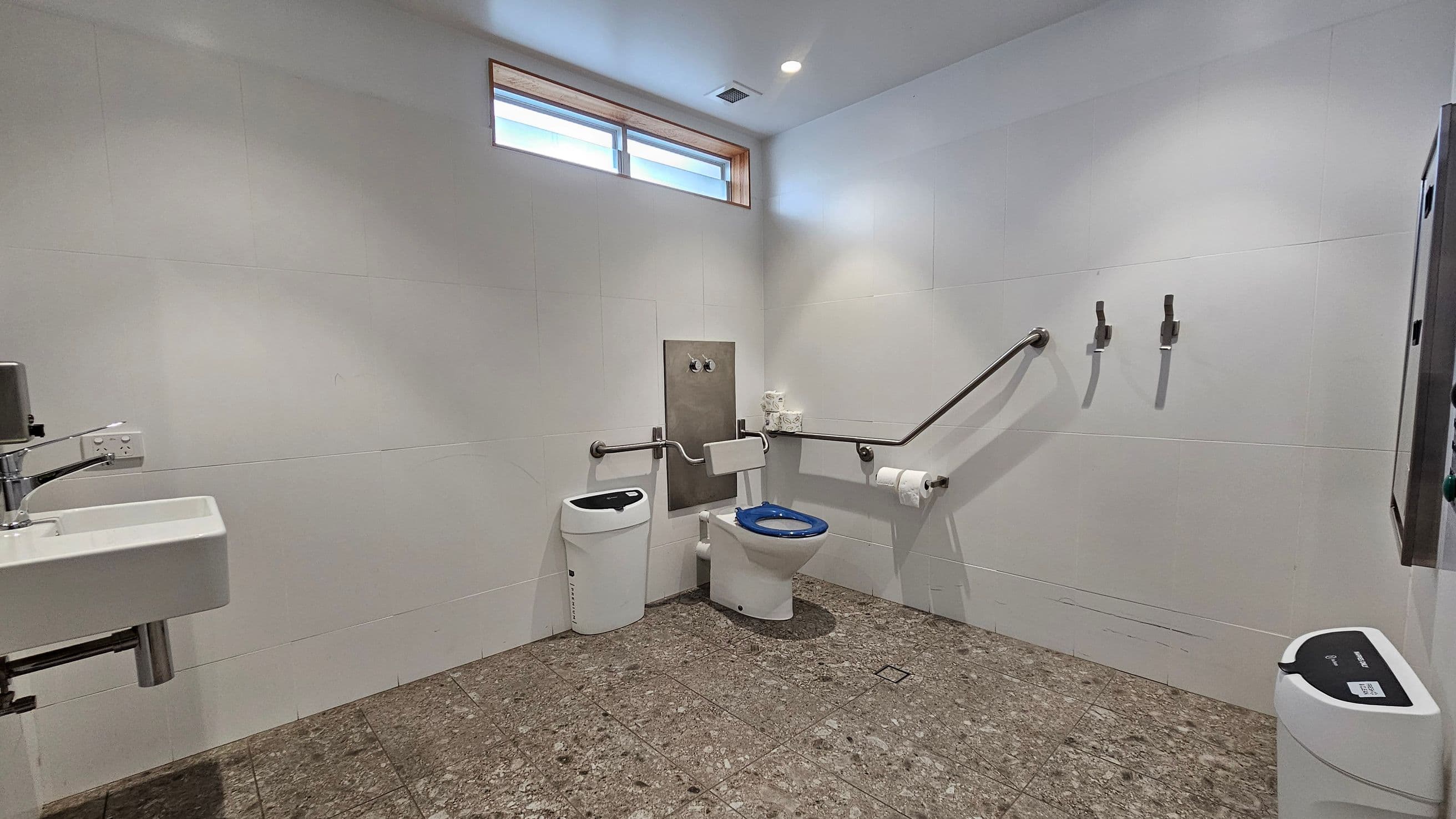 A spacious, accessible bathroom with a white toilet featuring a blue seat in the centre. To the left, a white sink with a silver tap is mounted on the wall. A small window near the ceiling allows natural light in. The floor is covered with brown tiles. On the right, a handrail and two hooks are attached to the wall, with a toilet roll holder nearby. A waste bin is positioned in the foreground on the right. The walls are plain and light-coloured.