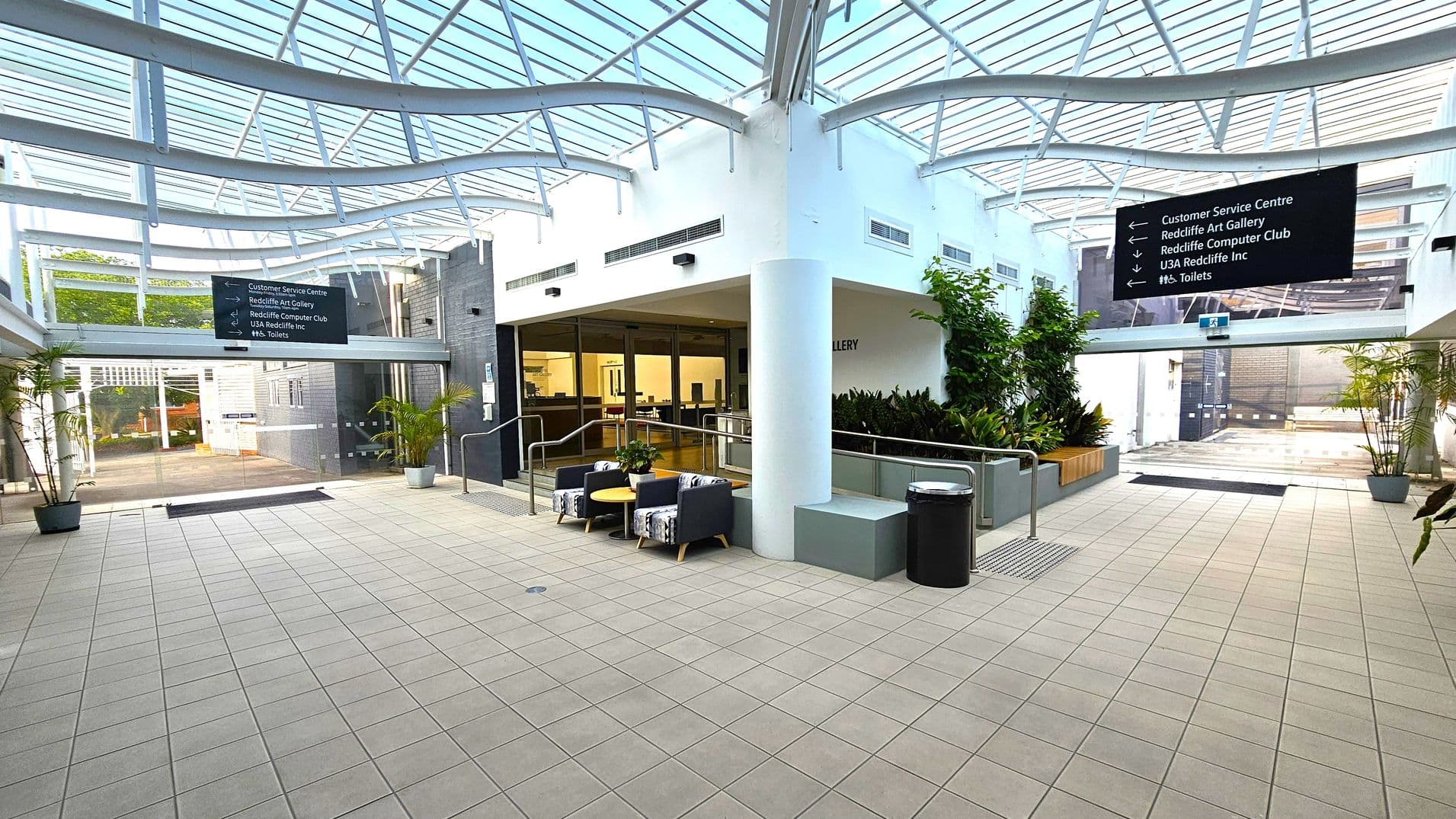 A spacious indoor area with a tiled floor and a glass ceiling supported by white beams. In the centre, there are two cushioned chairs and a small round table. To the right, a wall with a large window and a plant bed. On the left, a sign with directions to various facilities. Potted plants are placed around the space, and a black bin is near the seating area. The background shows a glass entrance with more plants outside.