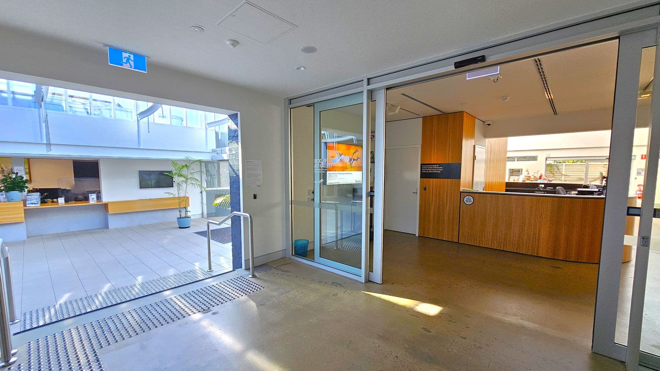 A modern indoor space with a polished concrete floor. On the right, there is a reception area with a wooden counter and a sign on the wall. A glass door separates this area from the rest of the space. On the left, an open area with a tiled floor features a long wooden bench with potted plants. Above, a blue exit sign is visible. The background shows large windows allowing natural light to illuminate the space.