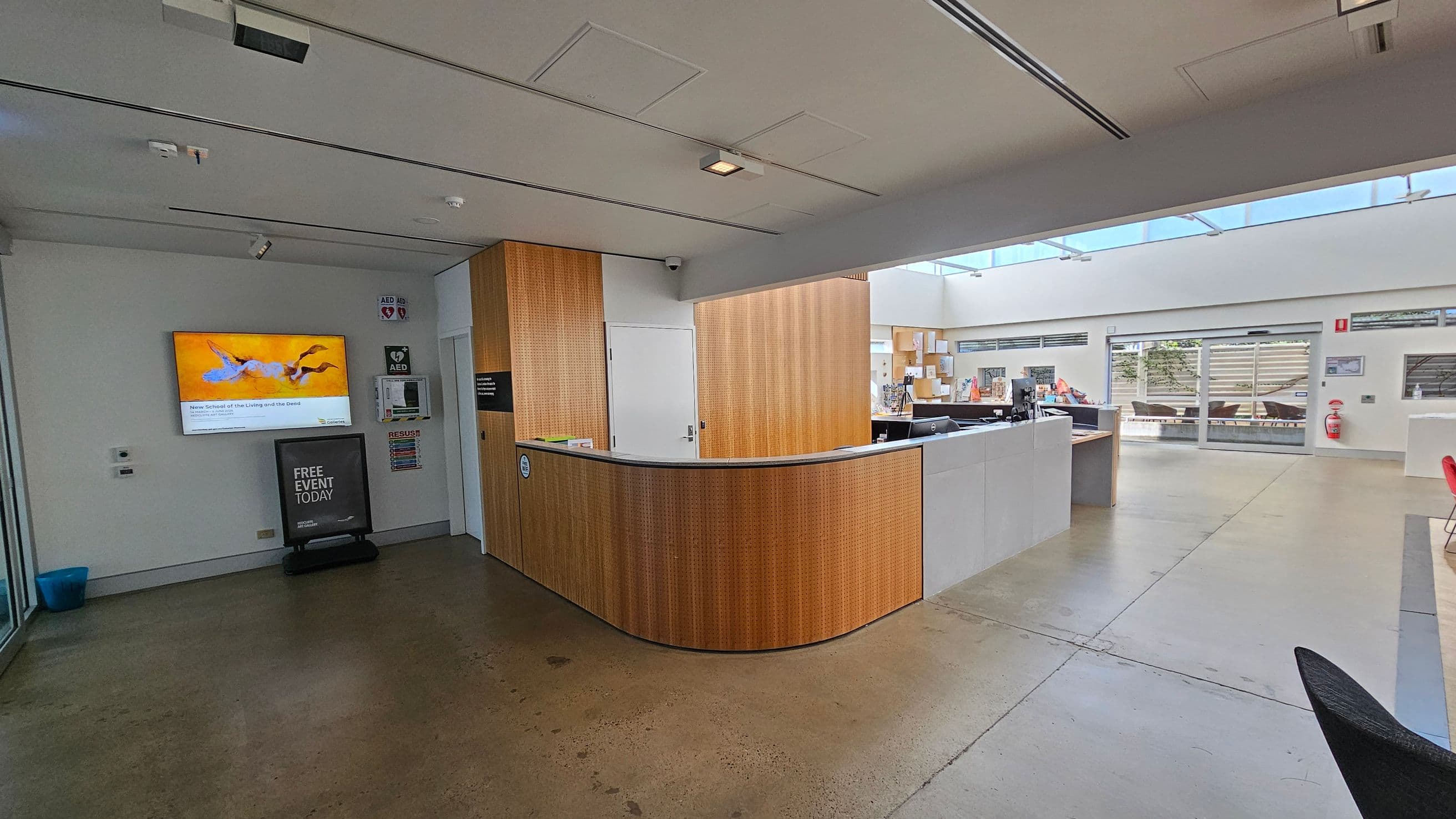 A modern interior space with a curved wooden reception desk in the centre. On the left, a wall-mounted screen displays an image with a bright yellow background, and below it is a sign reading "FREE EVENT TODAY." The floor is polished concrete. On the right, large windows and glass doors allow natural light to fill the room, with a view of outdoor seating. The ceiling is white with recessed lighting, and there are wooden panels on the walls behind the desk.
