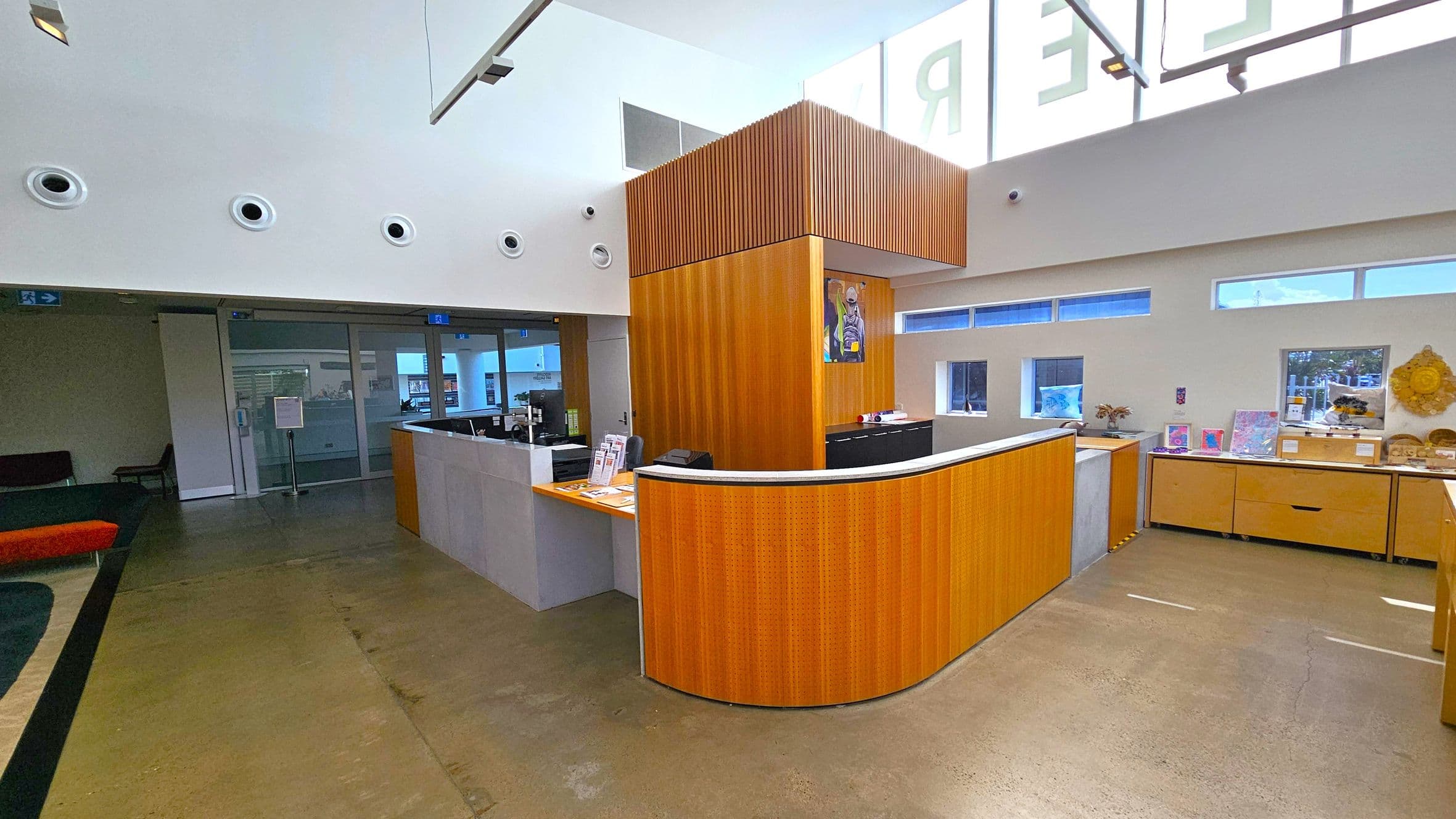 A modern reception area with a curved wooden desk in the centre. Behind the desk, a tall wooden panel extends to the ceiling. On the right, there are small windows and a display of artwork and decorative items on a cabinet. The left side features a glass door leading to another room, with a sign and a few chairs visible. The floor is polished concrete, and the ceiling is high with circular vents and large windows allowing natural light.