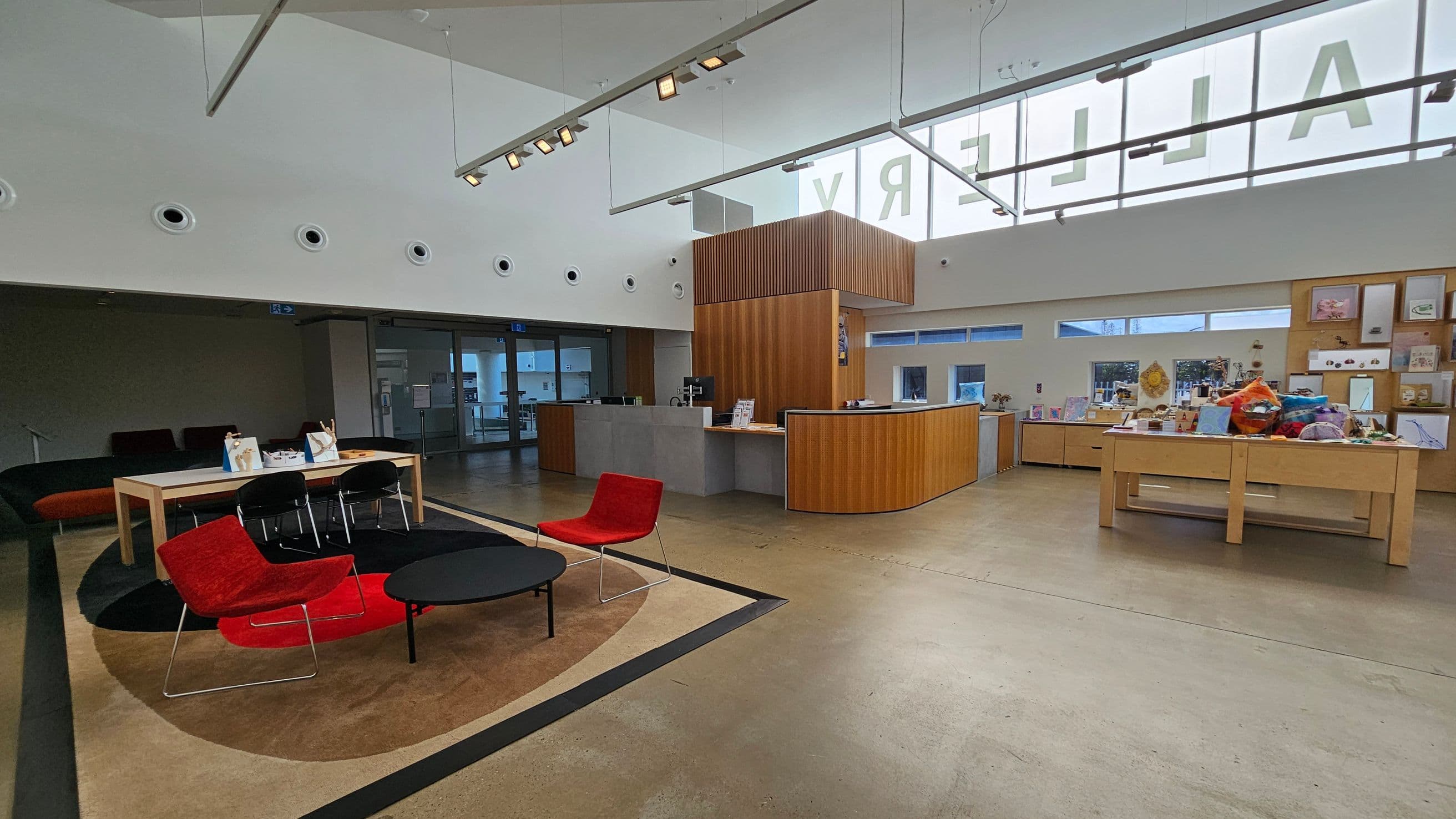 A spacious interior with a modern design. On the left, there is a seating area with red chairs and a black coffee table on a circular rug. A wooden table with black chairs is nearby. In the centre, a reception desk with a wooden and grey finish is positioned. On the right, shelves display various items, including books and decorative objects. The ceiling is high with circular lights and a large window above, partially showing letters. The floor is polished concrete.