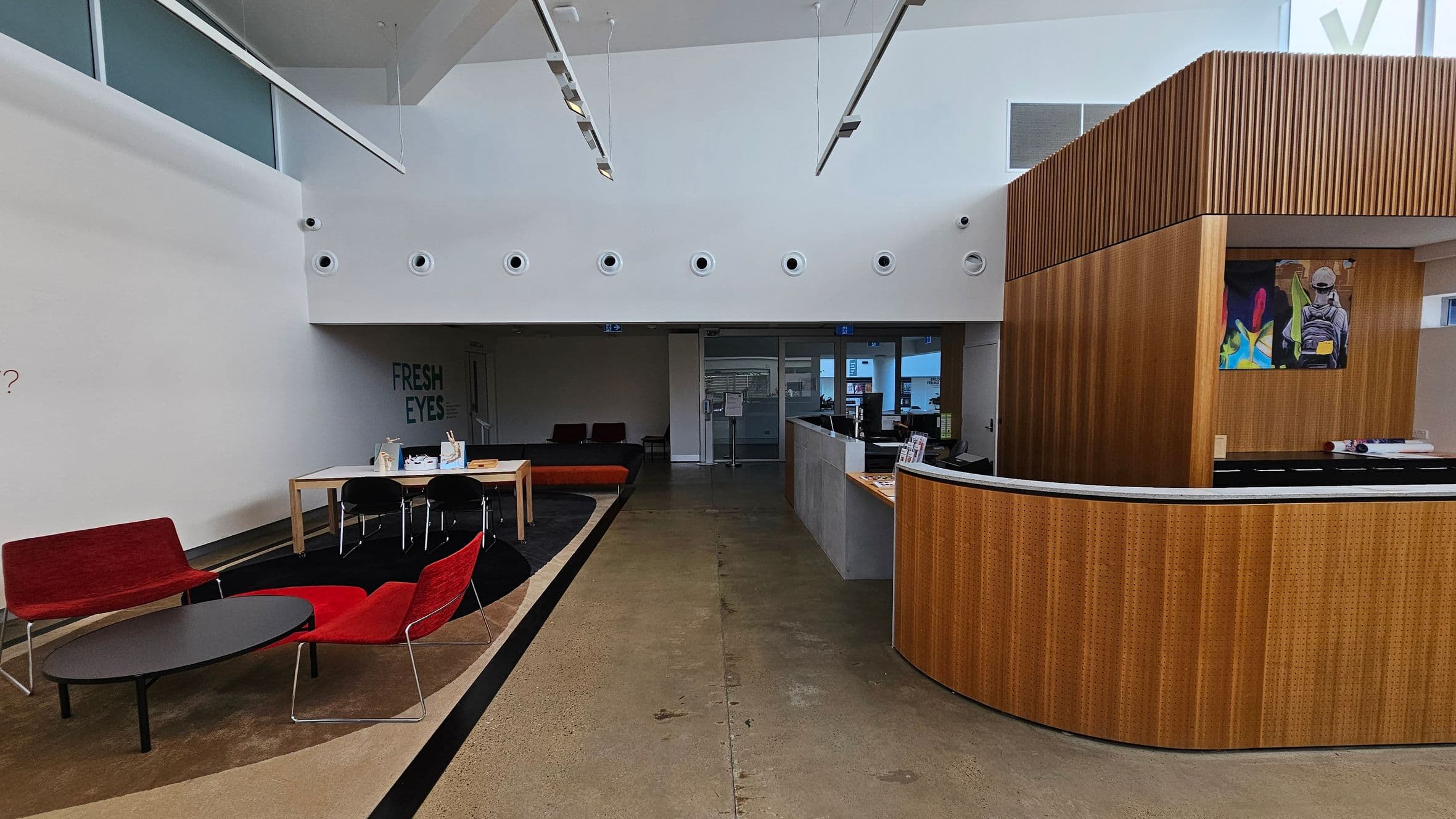 A modern interior space with a high ceiling. On the left, there are red chairs and a black oval table on a patterned carpet. A wooden table with black chairs is nearby. The back wall has the words "FRESH EYES". On the right, a curved wooden reception desk with a colourful artwork above it. The floor is a mix of concrete and carpet, and the ceiling features circular vents and hanging lights.