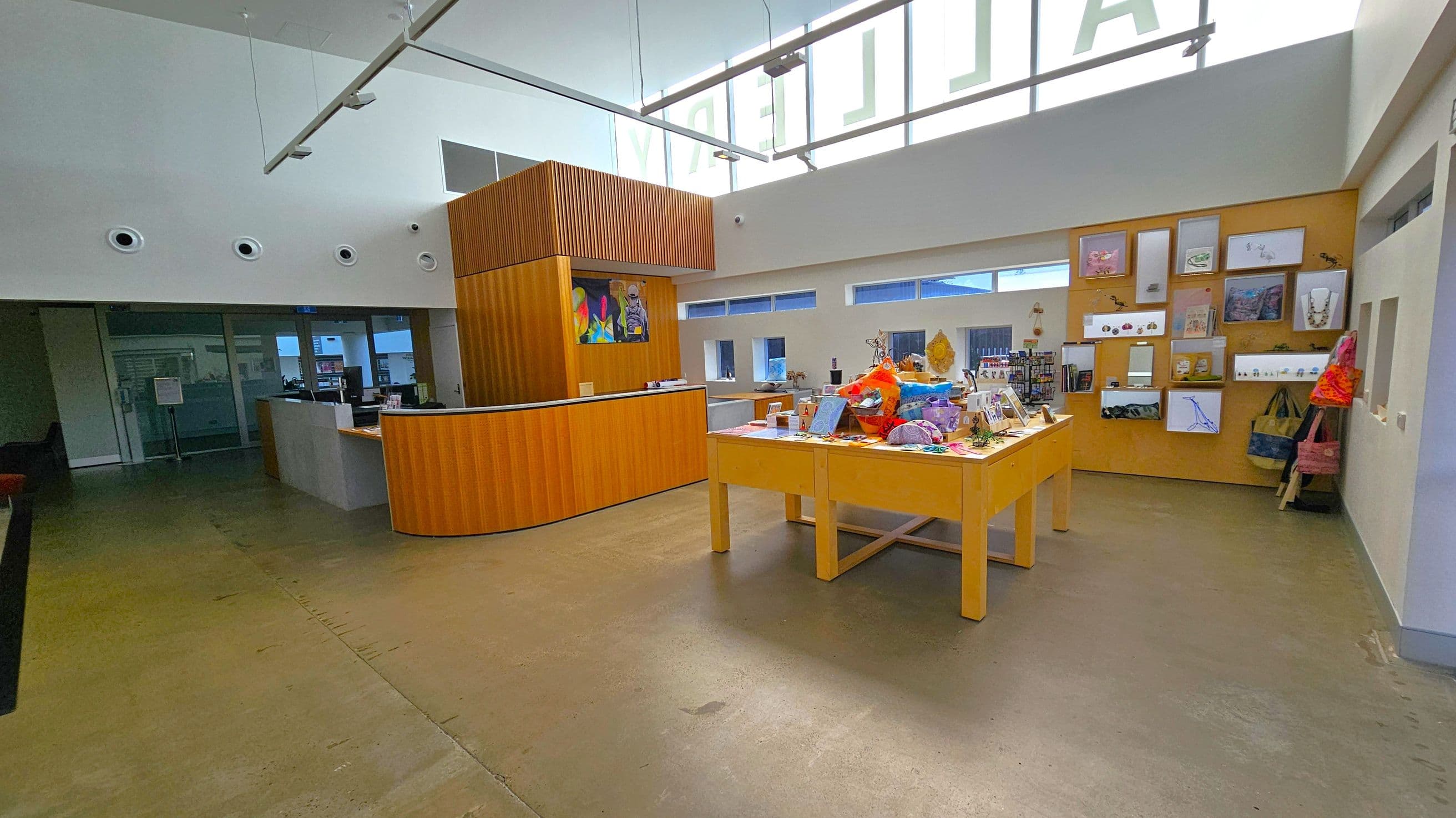 A spacious interior with a polished concrete floor. On the left, there's a curved wooden reception desk with a colourful artwork above it. In the centre, a wooden table displays various items, including crafts and books. The right wall features a display of framed pictures and hanging bags. The ceiling is high with modern lighting, and there are small windows along the back wall, allowing natural light to enter.