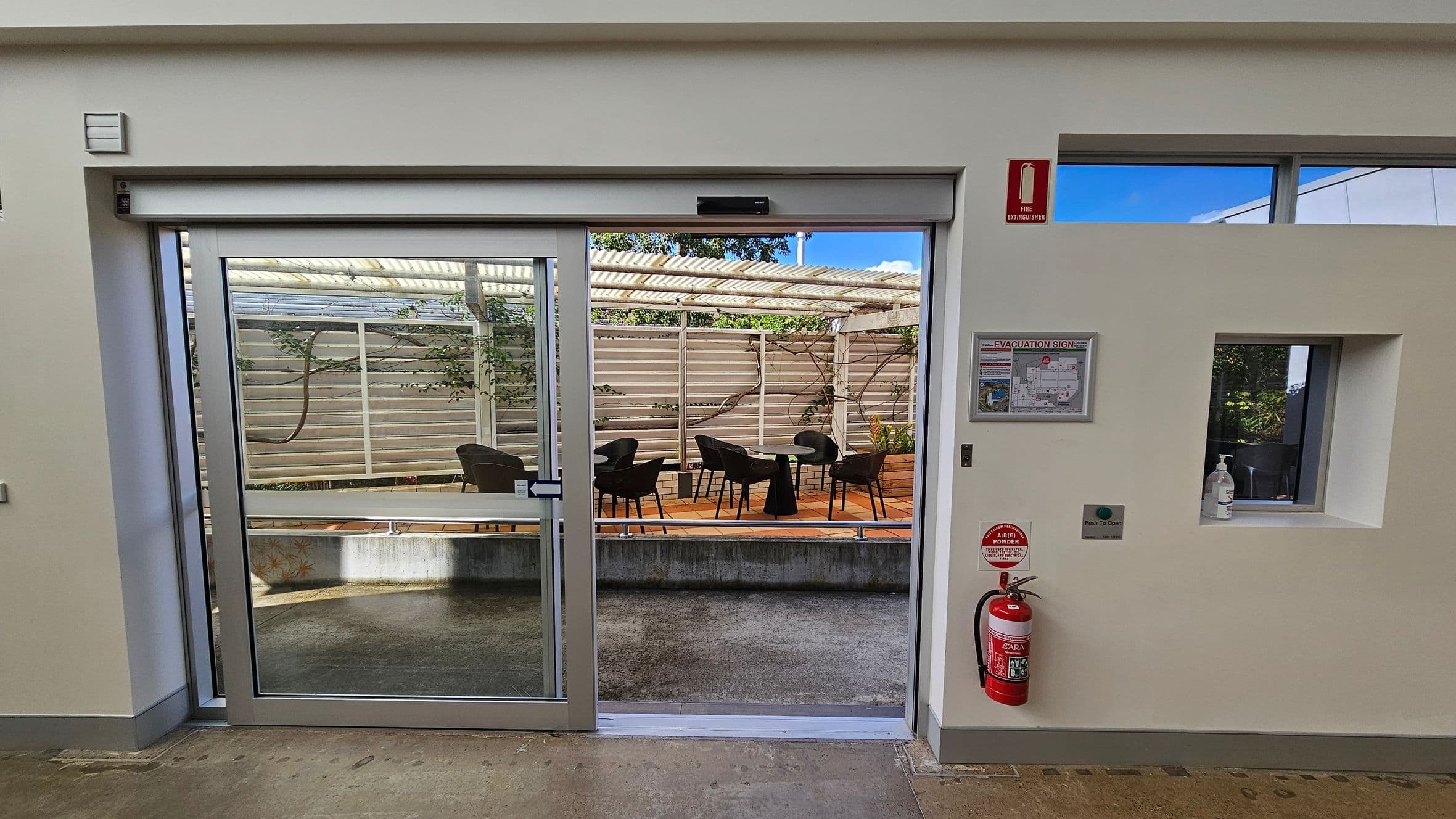 A glass sliding door leads to an outdoor patio with black chairs and tables on a terracotta-tiled floor. The patio is enclosed by a wooden fence with climbing plants. Inside, a fire extinguisher is mounted on the right wall below an evacuation sign. A small window with a hand sanitiser bottle is also on the right. The floor is a mix of concrete and tiles, and the ceiling is plain white.