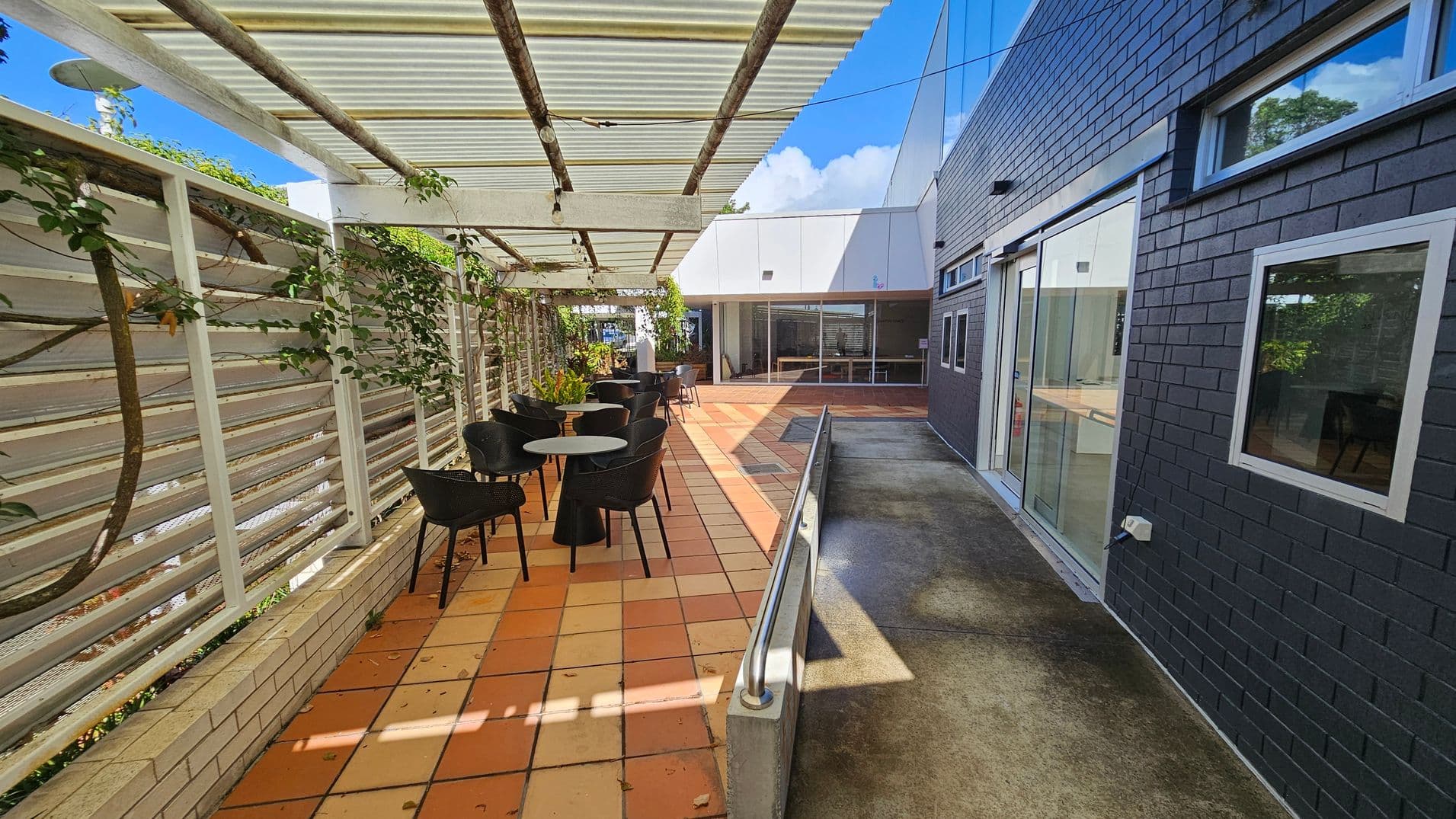 A covered outdoor patio area with terracotta tiles on the floor. On the left, a white trellis with climbing plants provides partial shade. Black chairs and round tables are arranged along the patio. On the right, a dark brick wall with windows and a sliding glass door leads to an indoor space. A metal handrail runs along a concrete path. In the background, a white building with large windows is visible. The ceiling is made of corrugated material with wooden beams.