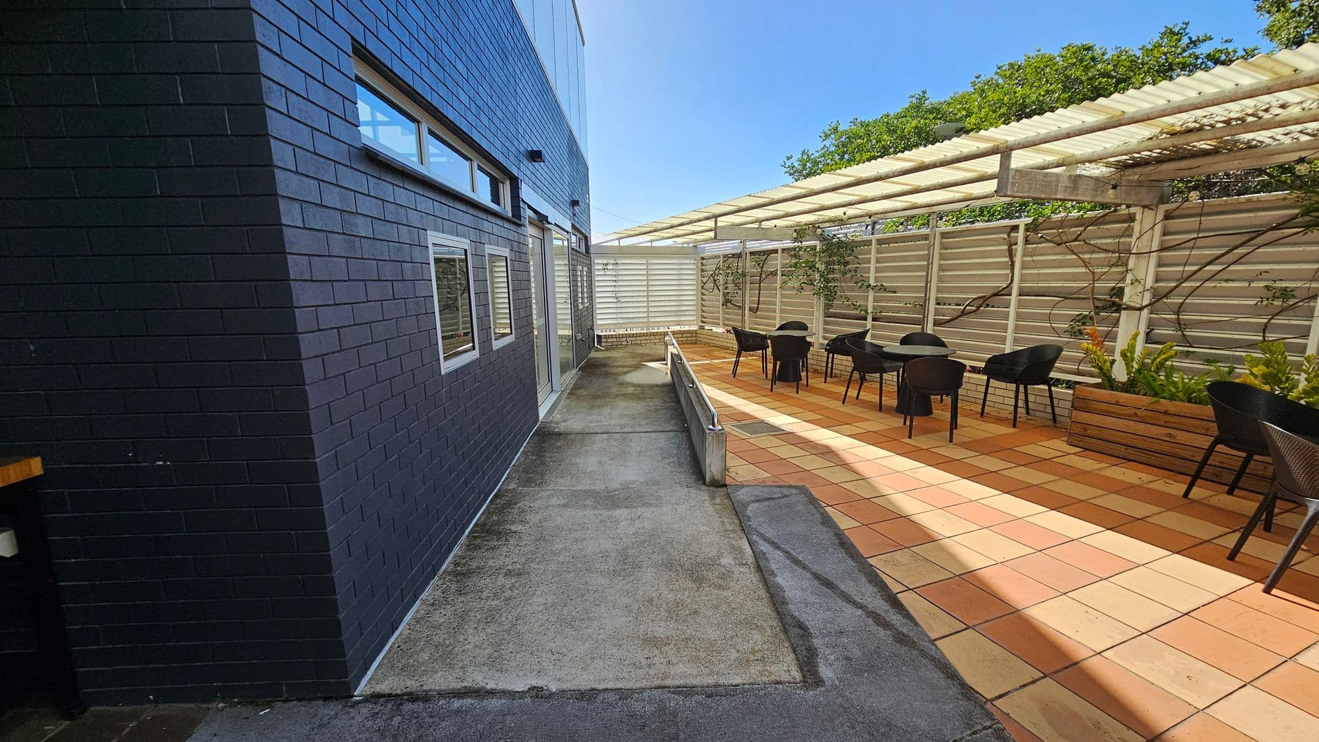 On the left, a dark brick wall with several windows and a door. The ground is a mix of concrete and moss. On the right, a patio area with terracotta tiles, featuring black chairs and tables. A pergola with a translucent roof provides shade, and there are wooden planters with green plants along the fence. The background shows a tall fence with climbing plants.