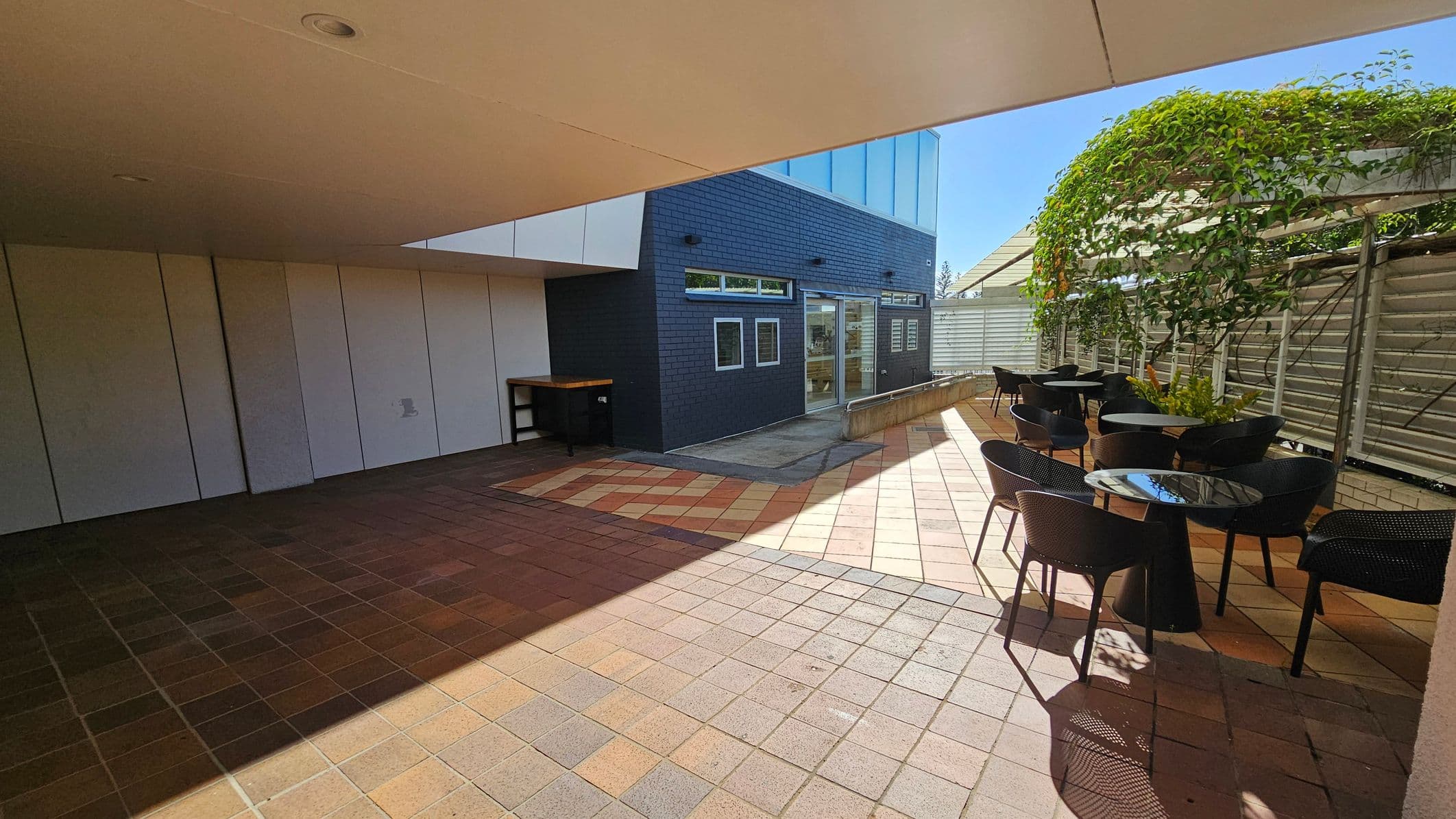 A covered outdoor area with a tiled floor. On the left, there is a wall with white panels and a small table. In the centre, a dark brick wall with several windows and a glass door. On the right, a seating area with black tables and chairs, partially shaded by a pergola with green vines. The ceiling is smooth and light-coloured, with a recessed light fixture.