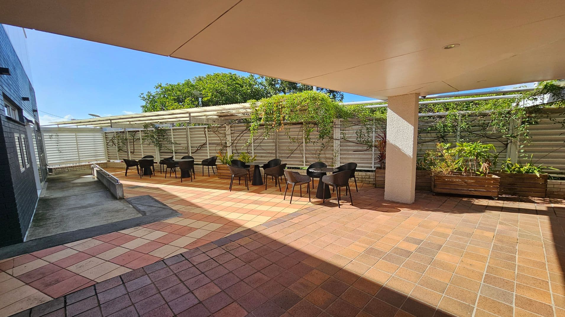 A spacious outdoor patio area with terracotta-coloured tiles. On the right, there are several black chairs and small round tables arranged in a row. Behind them, a trellis with climbing plants and greenery adds a natural touch. On the left, a dark brick wall and a concrete path lead to the seating area. A large white ceiling overhang provides shade, and a white pillar supports it. The background features more greenery and a clear blue sky.
