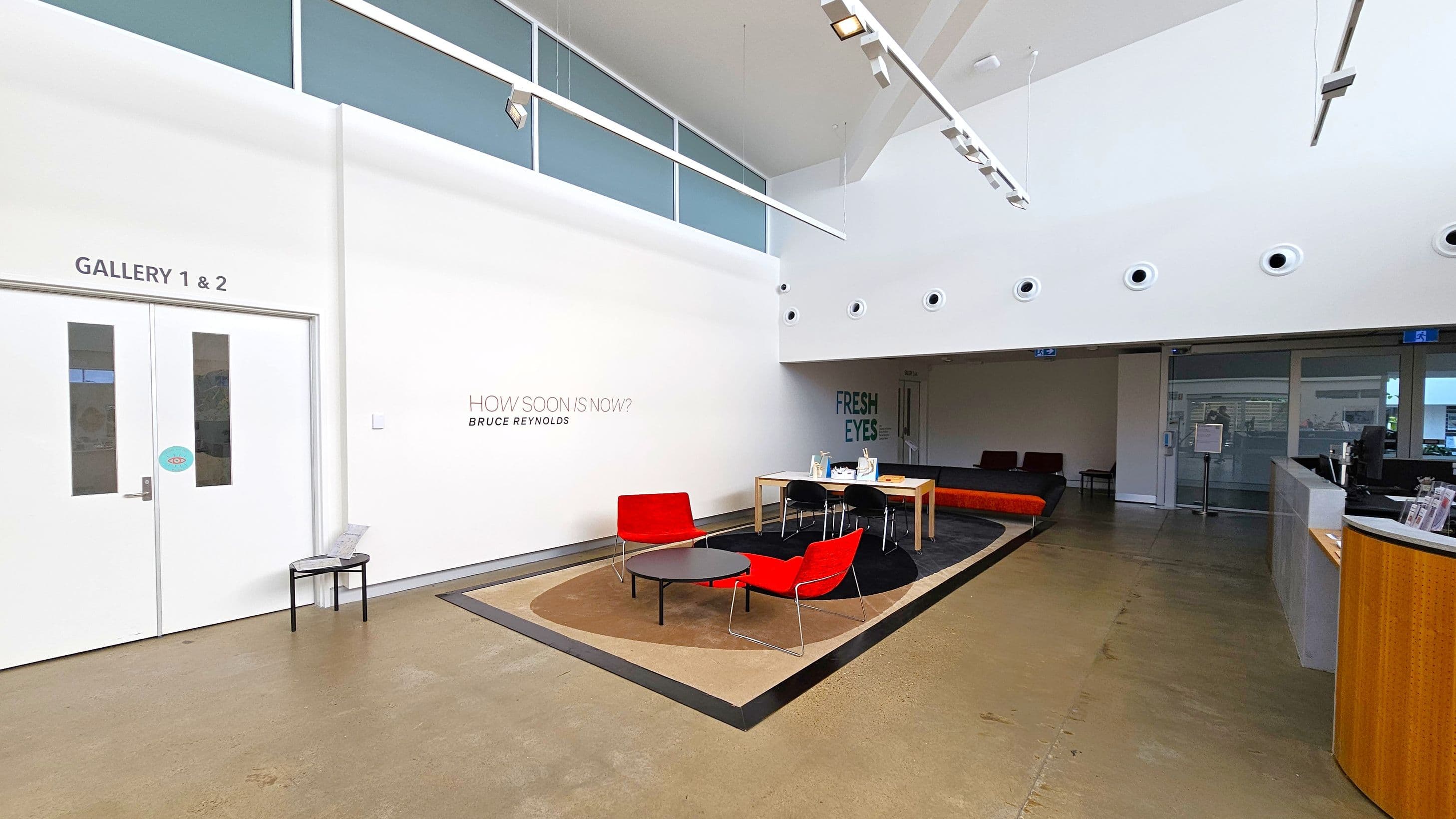 A modern interior with a polished concrete floor. On the left, double doors labelled "Gallery 1 & 2" are next to a small black table. The centre features a seating area with two red chairs and a round black table on a beige and black rug. A wooden table with black chairs is nearby. The right side has a reception desk with brochures. The back wall displays the text "HOW SOON IS NOW? BRUCE REYNOLDS" and "FRESH EYES". The ceiling is high with circular vents and track lighting.