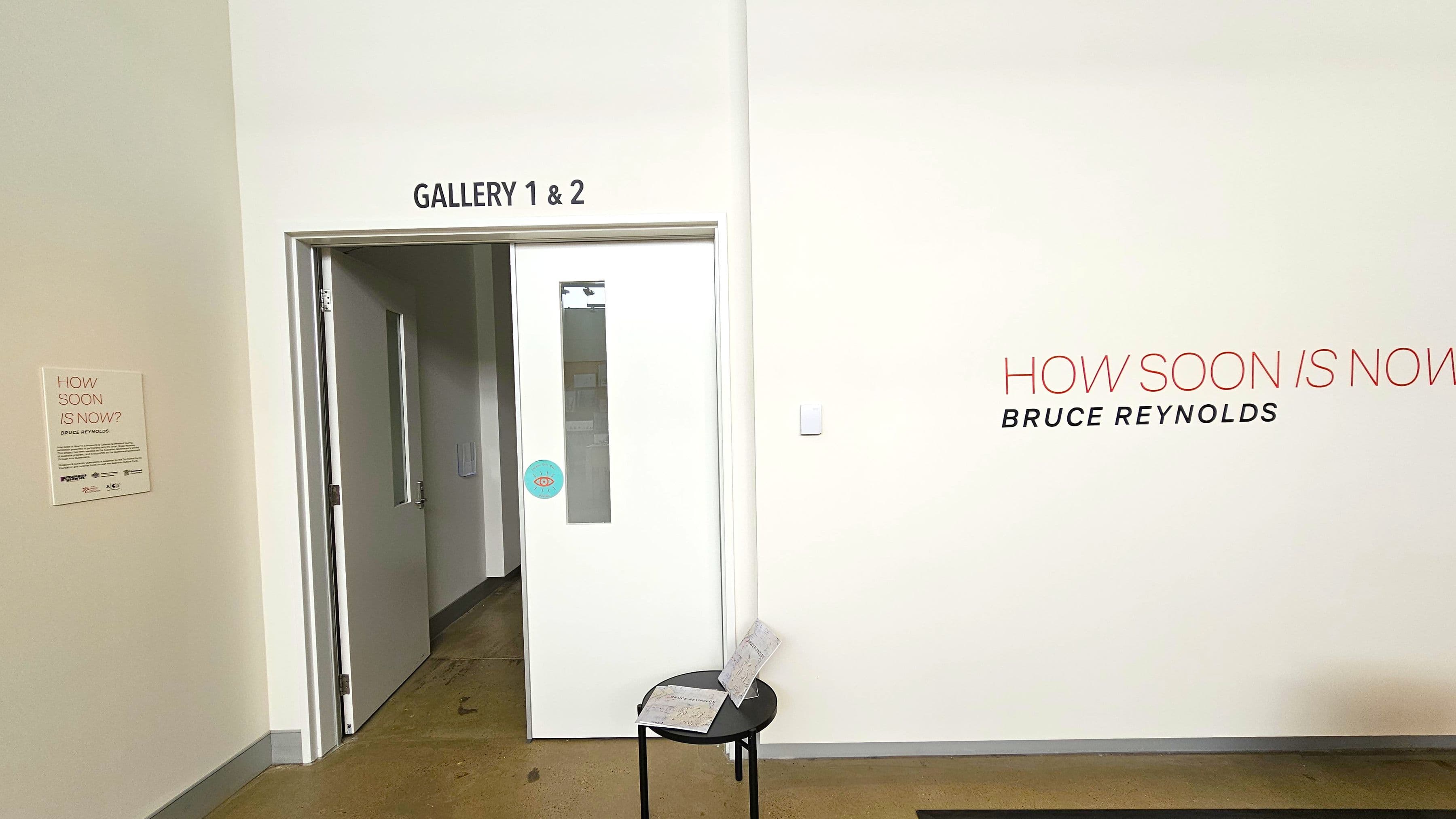 A white wall with the text "GALLERY 1 & 2" above a partially open door on the left. Next to the door, a small sign reads "HOW SOON IS NOW?" with additional text below. On the right, large text on the wall says "HOW SOON IS NOW" with "BRUCE REYNOLDS" underneath. A small black table in front of the wall holds some papers. The floor is a polished concrete surface.