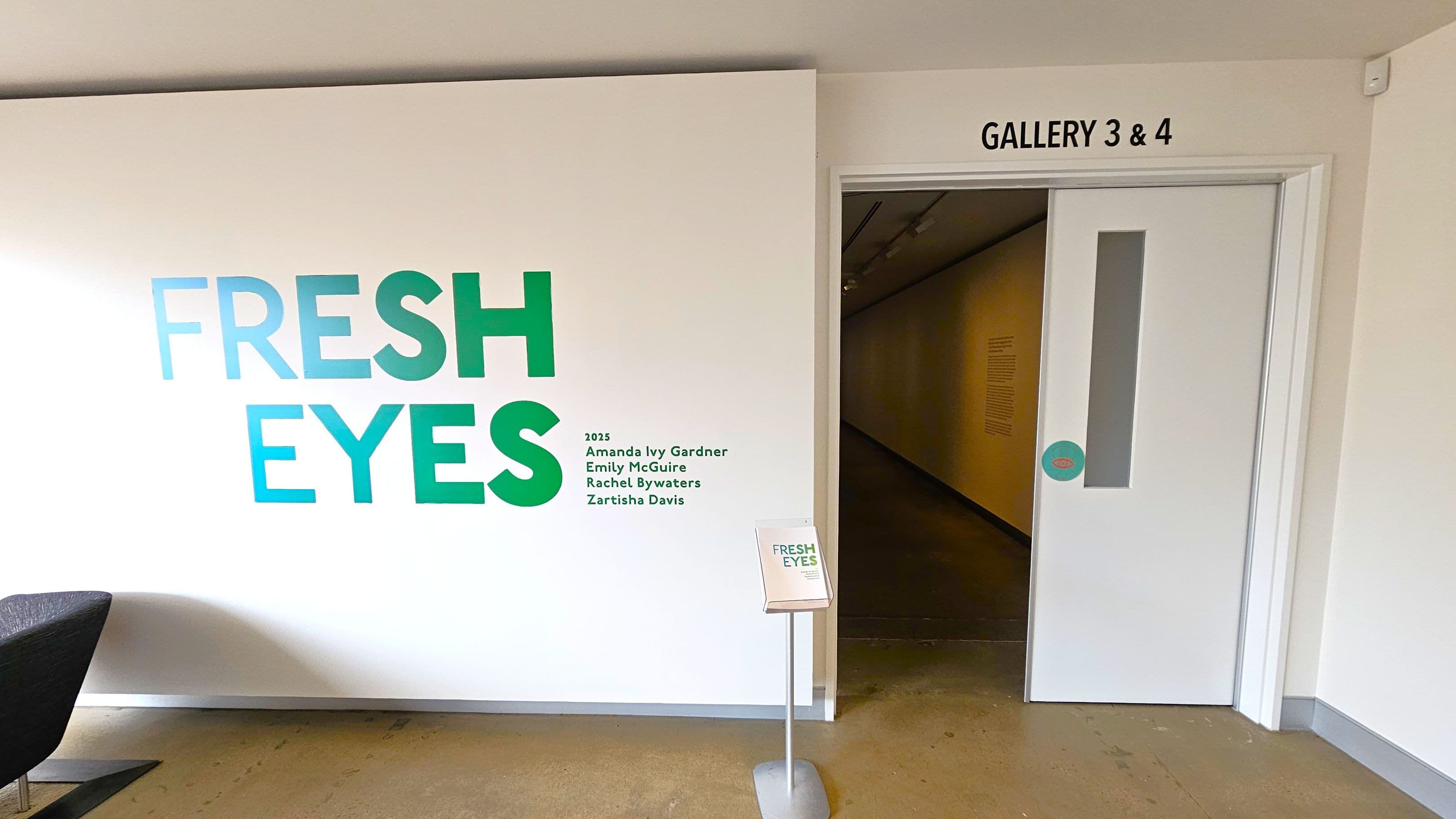 On the left, a white wall displays the text "FRESH EYES" in large green and blue letters, with names listed below. A small stand with a sign also reads "FRESH EYES." On the right, a partially open white door leads to a hallway, marked "GALLERY 3 & 4" above. The floor is a polished concrete surface.
