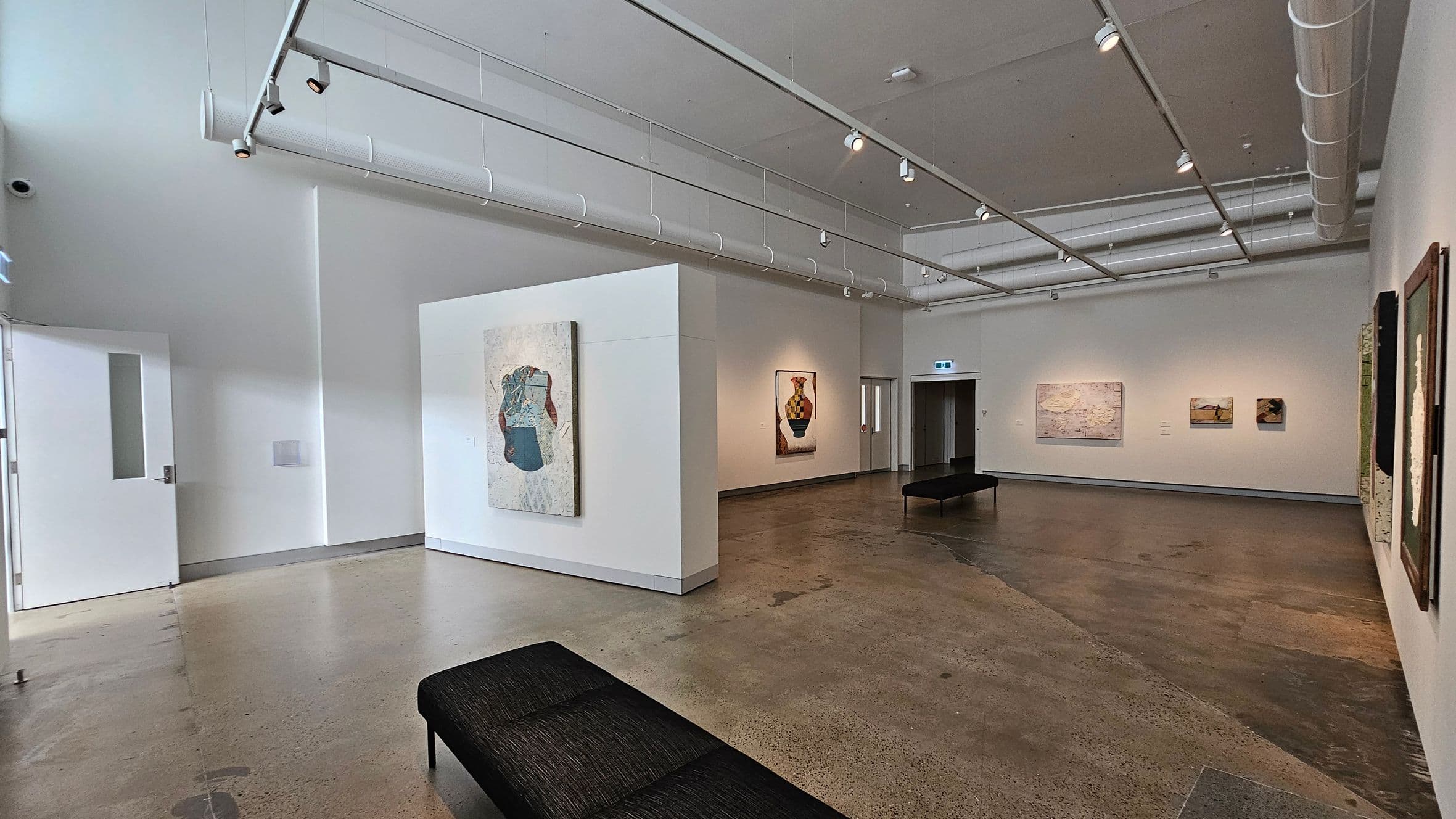 A modern art gallery with polished concrete floors and a high ceiling featuring exposed ductwork and track lighting. On the left, a white wall with a door and a large abstract painting. In the centre, a partition wall displays another abstract artwork. The right side has several paintings, including a prominent one of a vase. Two black benches are placed in the middle of the room. The gallery is well-lit, highlighting the artworks.