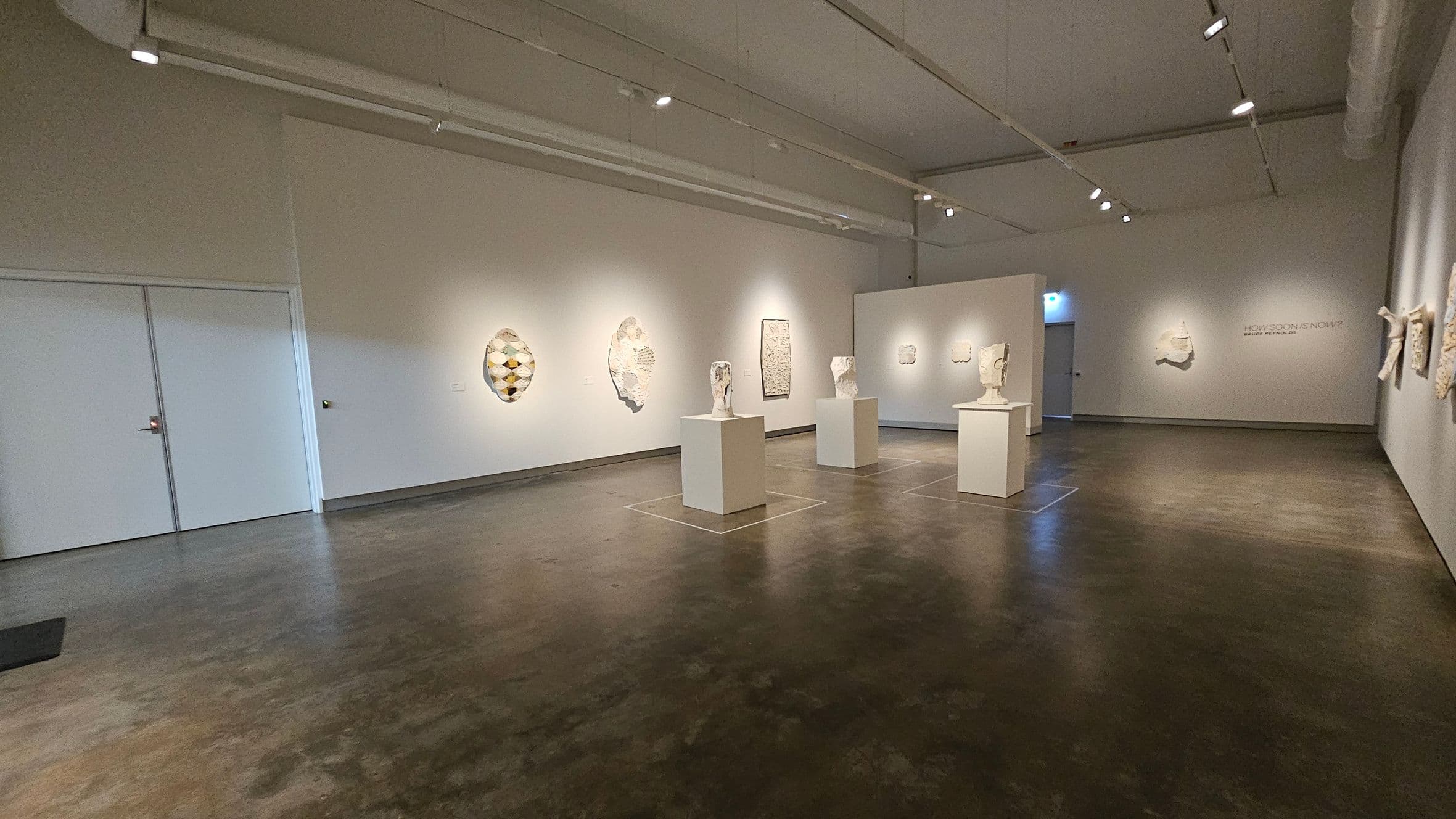 A spacious art gallery with a polished concrete floor and a high ceiling with track lighting. In the centre, several white sculptures are displayed on square pedestals. The left wall features abstract wall art, while the right wall has more sculptures mounted. At the back, a doorway is visible, and the words "HOW SOON IS NOW?" are written on the wall. The gallery is well-lit, highlighting the artworks.