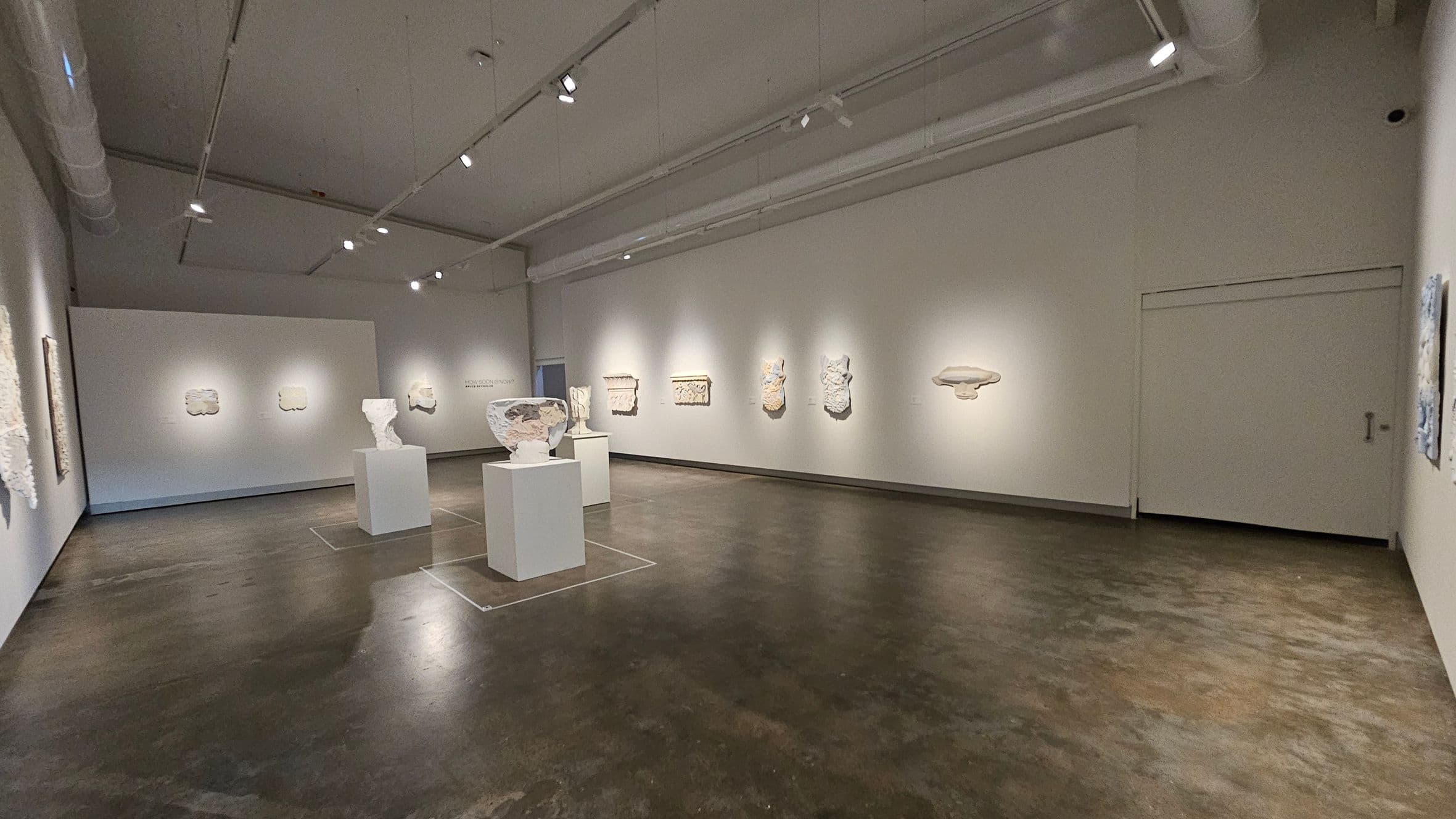 A spacious art gallery with a polished dark floor and a high ceiling featuring exposed ducts and track lighting. On the left and centre, several sculptures are displayed on white pedestals. The walls are adorned with various relief artworks, evenly spaced and lit by spotlights. The right side has a plain wall with a closed door. The overall atmosphere is minimalist and well-lit, highlighting the art pieces.
