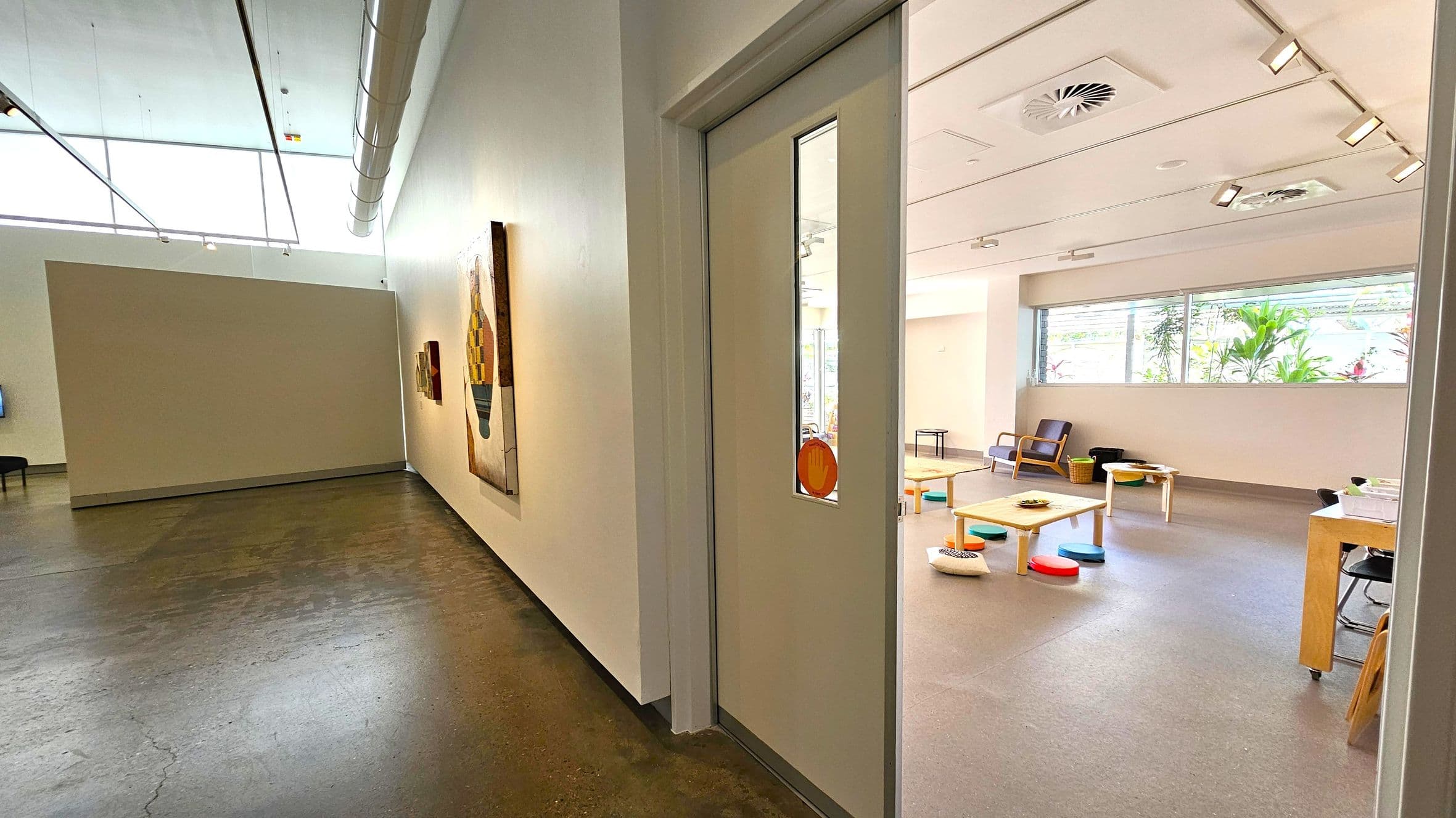 A hallway with a polished concrete floor on the left, featuring a large painting on the wall. On the right, a door with a narrow window opens into a room with a grey floor. Inside, there are low tables with colourful cushions and a few chairs. Large windows let in natural light, and there are plants visible outside. The ceiling has recessed lighting and air vents.