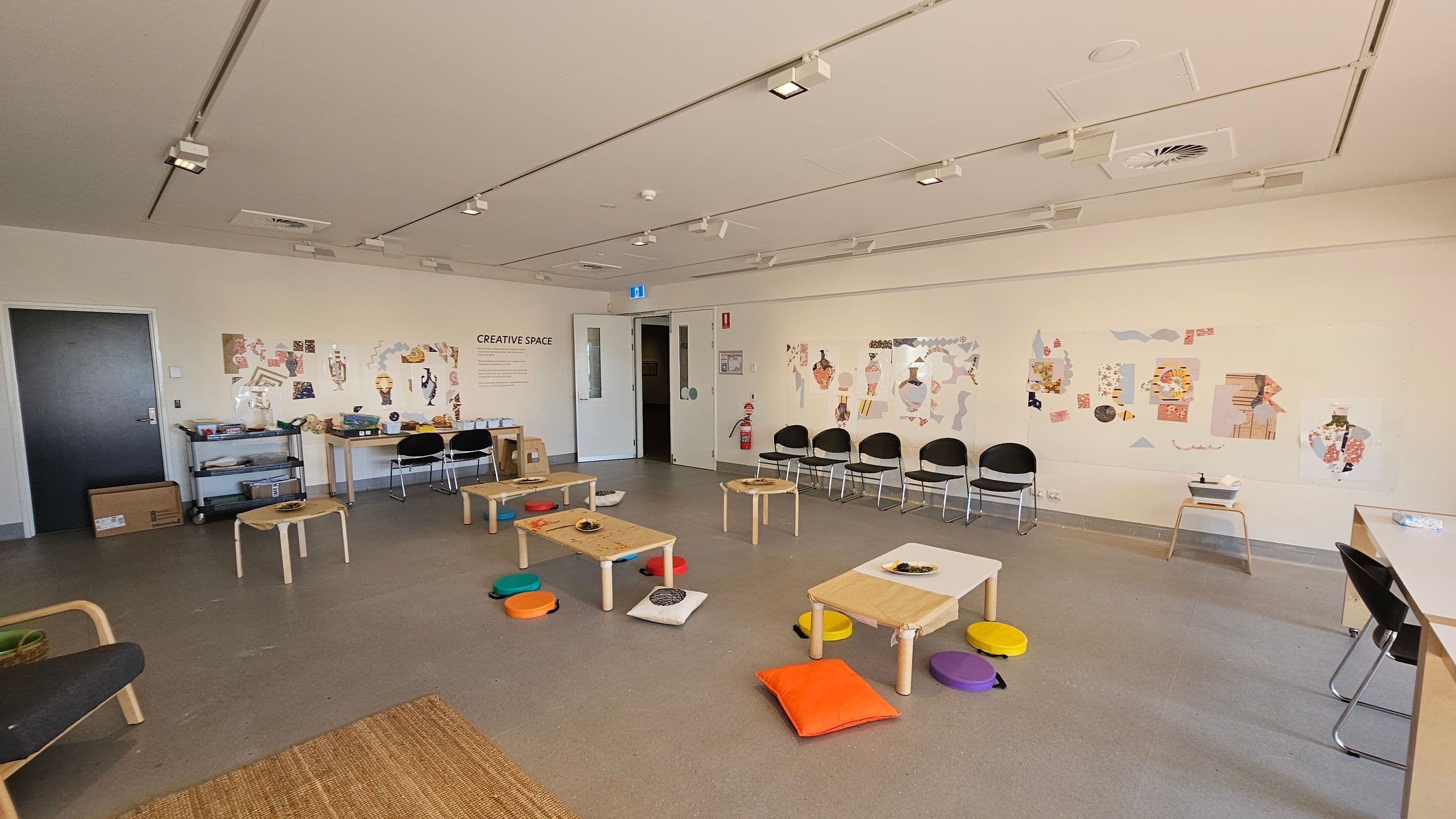 A spacious room with a light grey floor and a white ceiling featuring track lighting. On the left, a dark door is next to a shelf with art supplies and a cardboard box. In the centre, low wooden tables are surrounded by colourful cushions. The right side has a row of black chairs against the wall, which is decorated with various art pieces. A sign reads "CREATIVE SPACE." A table with chairs is on the far right. The room has a clean, organised appearance.
