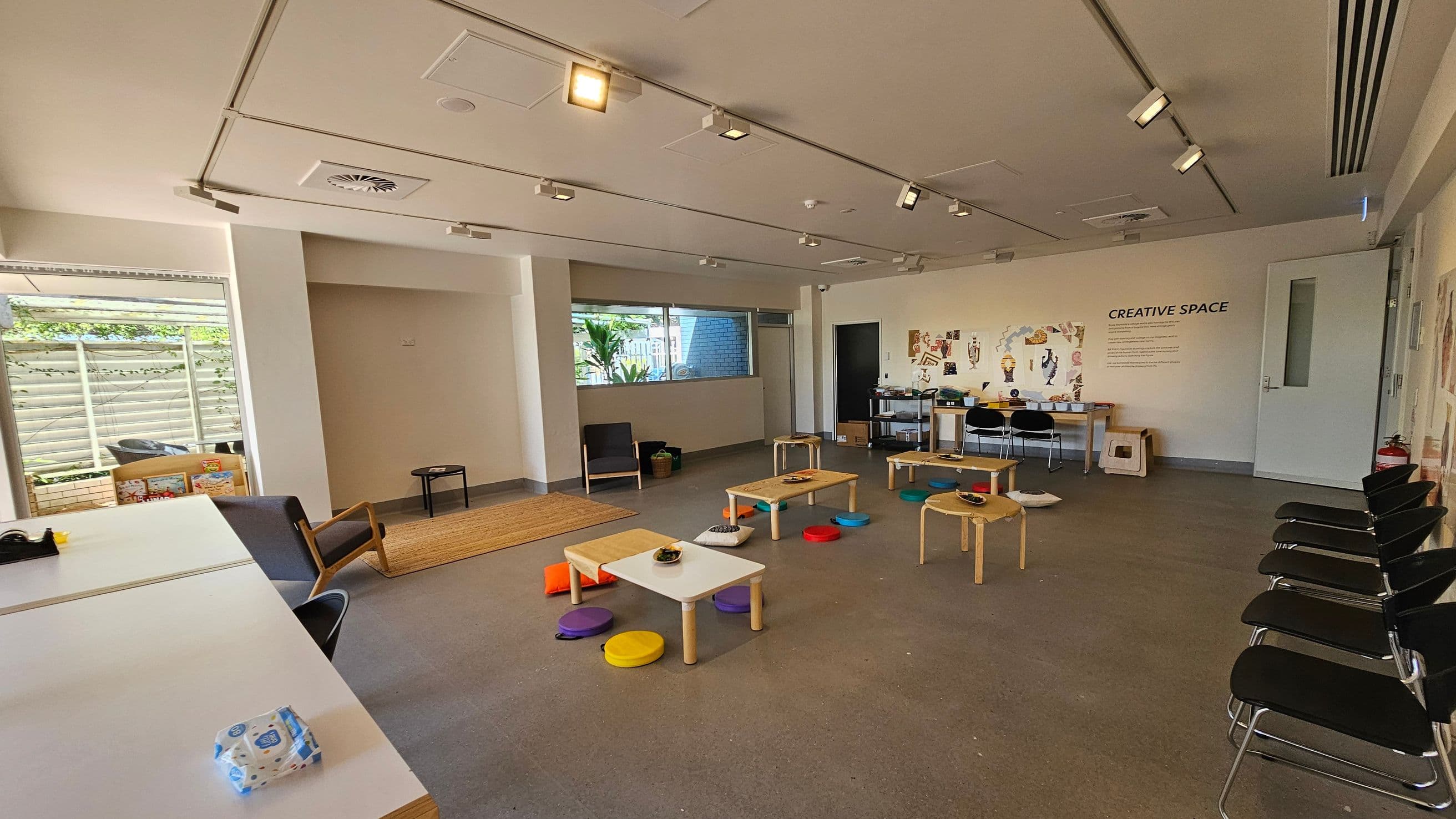 A spacious room with a creative theme. On the left, there's a large window with a view of greenery outside, and a few children's books are stacked nearby. In the centre, low tables are surrounded by colourful cushions on the floor. The right side features a wall with the words "CREATIVE SPACE" and various artworks displayed. Several chairs are lined up against the wall. The ceiling has track lighting, and the floor is a smooth, grey surface.