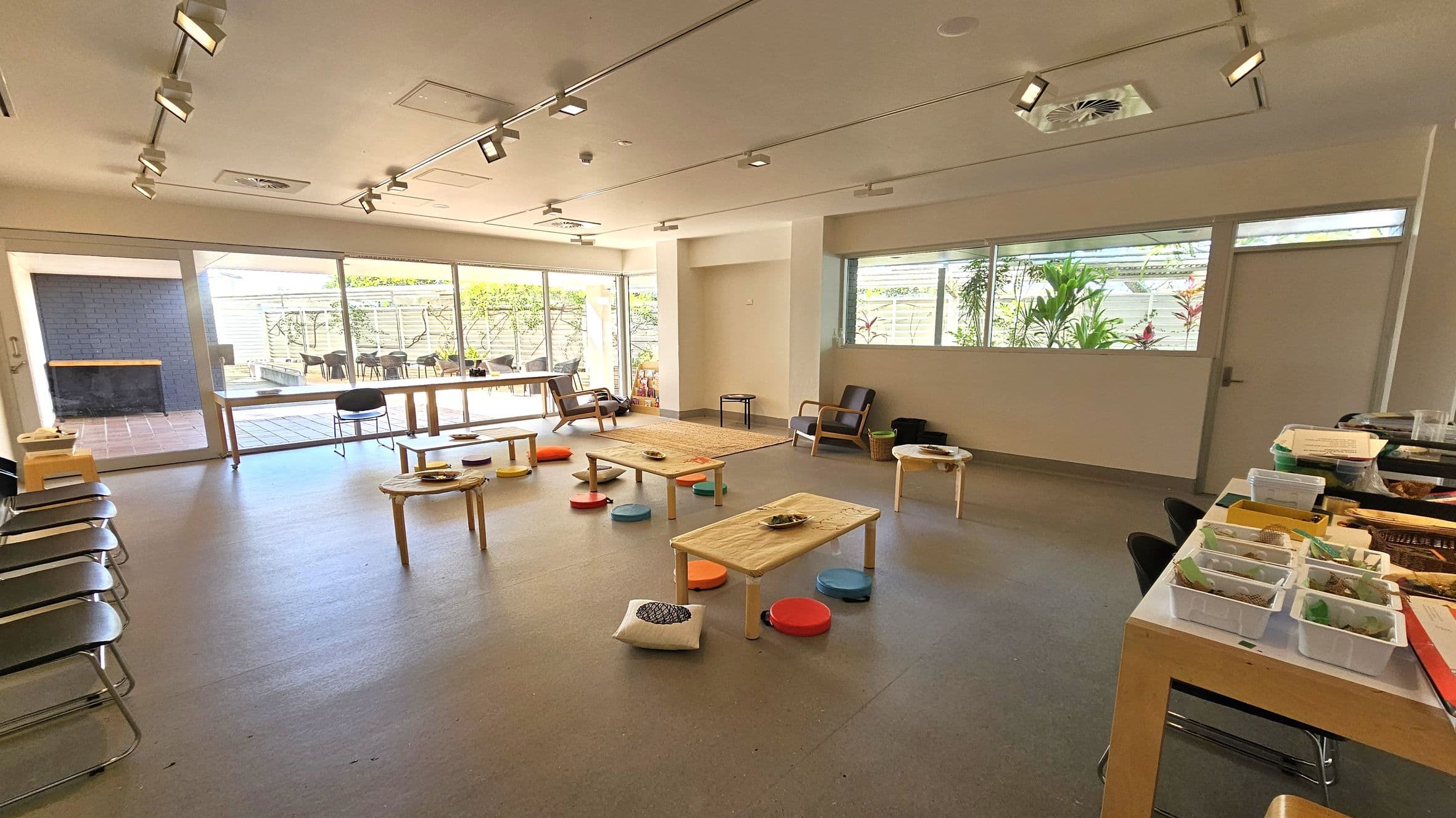 A spacious room with a light grey floor and a ceiling fitted with track lighting. In the centre, there are low wooden tables surrounded by colourful round cushions and a few square ones. On the right, a table holds various containers and materials. The left side features a row of chairs and large windows leading to an outdoor area with more seating. The back wall has windows with plants visible outside, and a few armchairs are placed against the walls.
