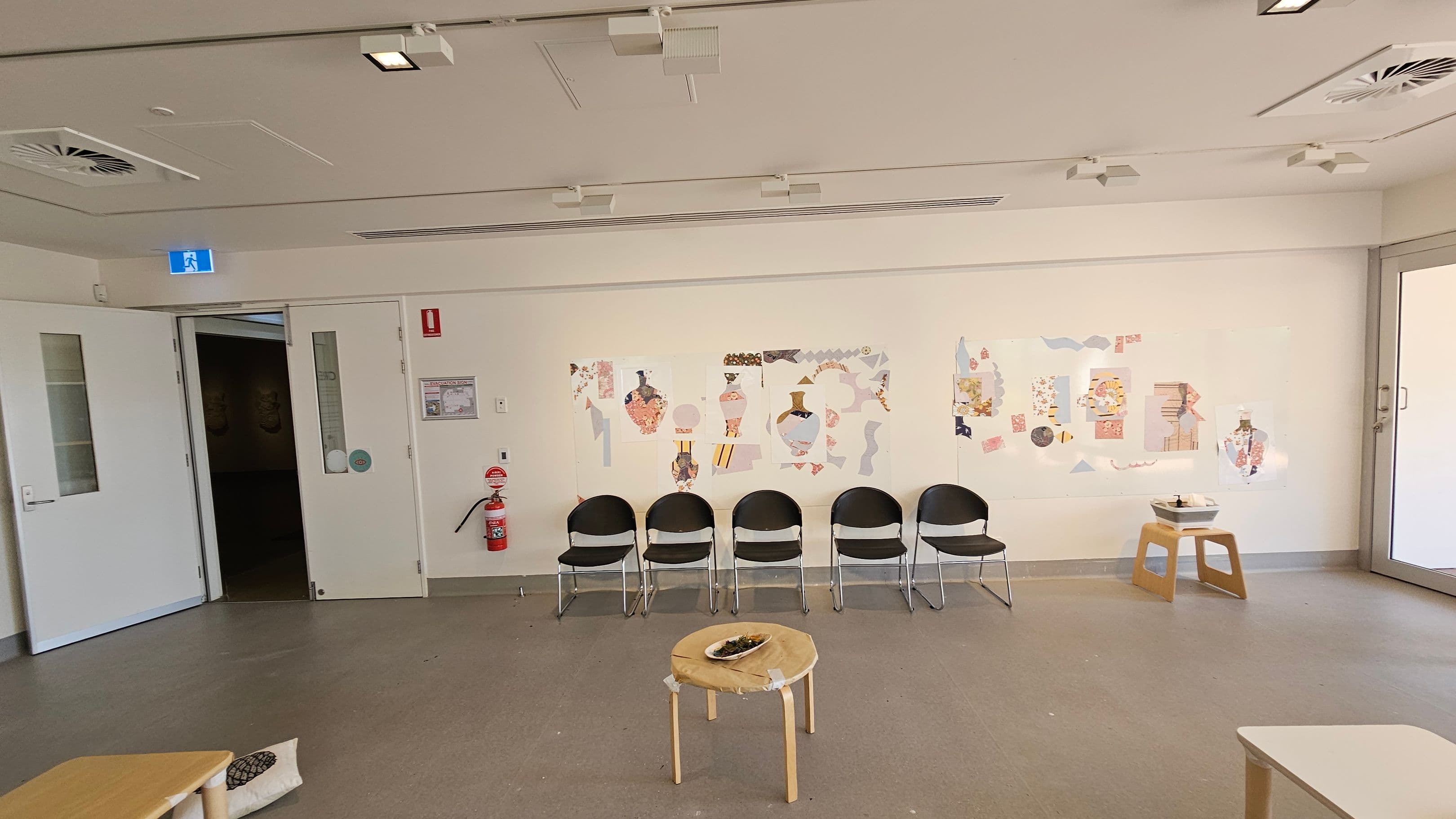 A room with a light-coloured ceiling featuring recessed lighting and air vents. On the left, there are double doors, one slightly open, with a fire extinguisher nearby. The wall displays colourful abstract art. In front of the art, six black chairs are lined up. A small round table with a dish is in the centre. On the right, a wooden stool holds a tray. The floor is grey and smooth, and a large window is on the far right.