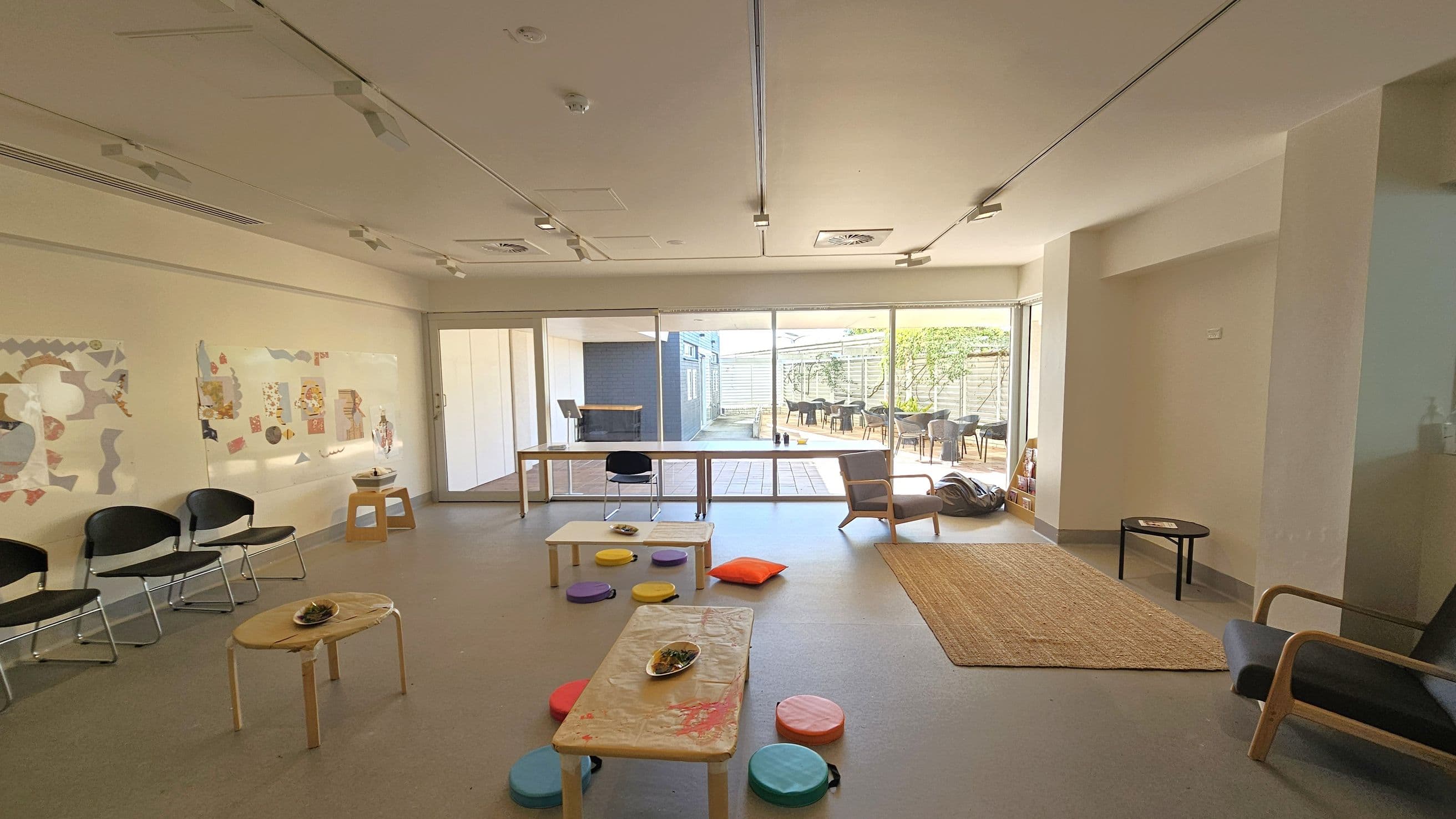 A spacious room with a light-coloured floor and ceiling. On the left, there are black chairs against the wall, which is decorated with colourful artwork. In the centre, low tables with colourful round cushions are scattered, and a few small dishes are placed on the tables. The right side has a woven rug and a wooden chair. Large glass doors at the back open to an outdoor area with more seating and greenery. The ceiling has multiple light fixtures.
