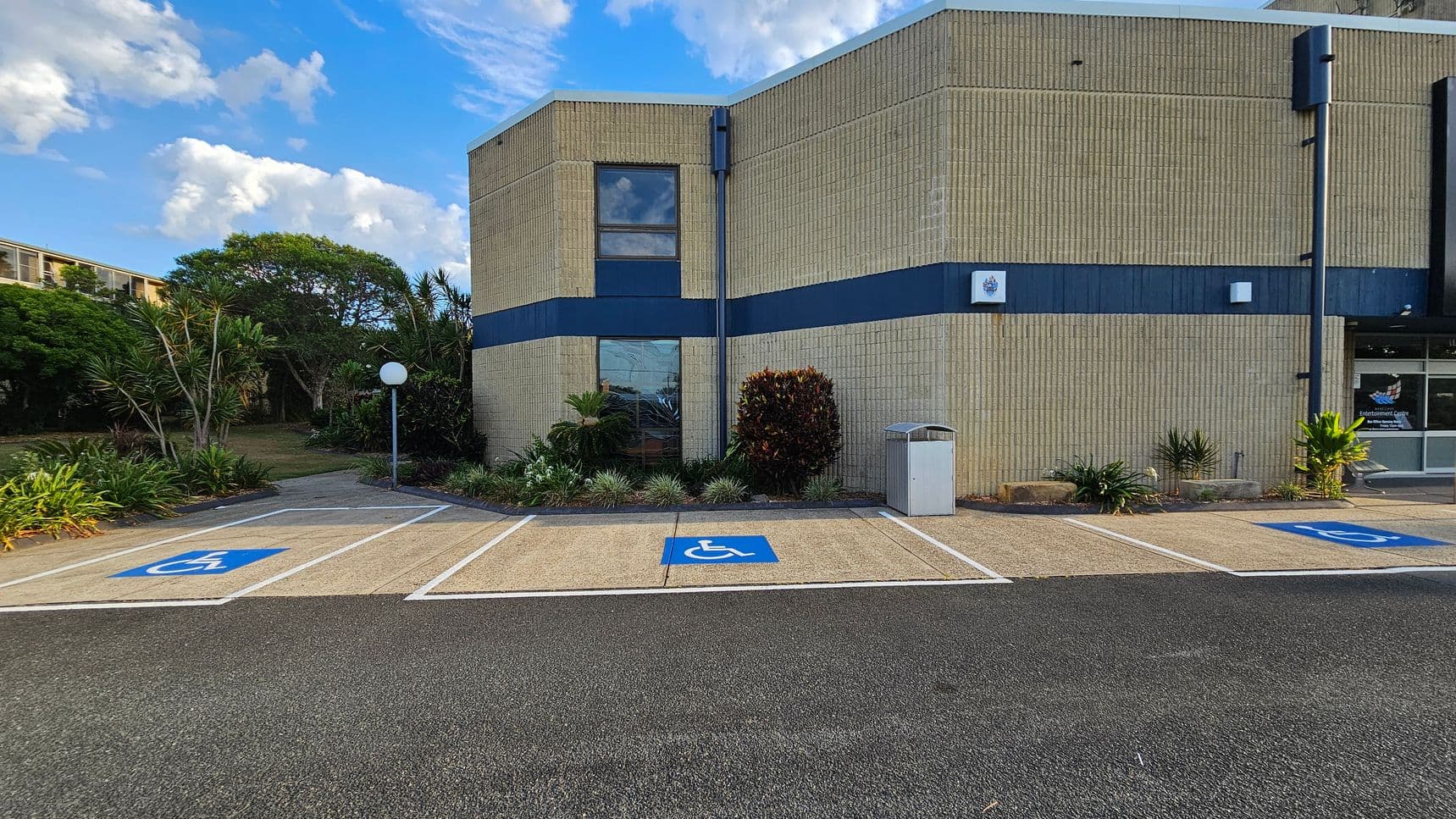 A building with light-coloured brick walls and a dark blue horizontal stripe. In front, there are three parking spaces marked with blue disabled signs. A small garden with various plants and a white spherical lamp post is on the left. A rubbish bin is positioned near the building. The entrance to the building is on the right, with glass doors and a sign. The ground is a mix of asphalt and concrete.